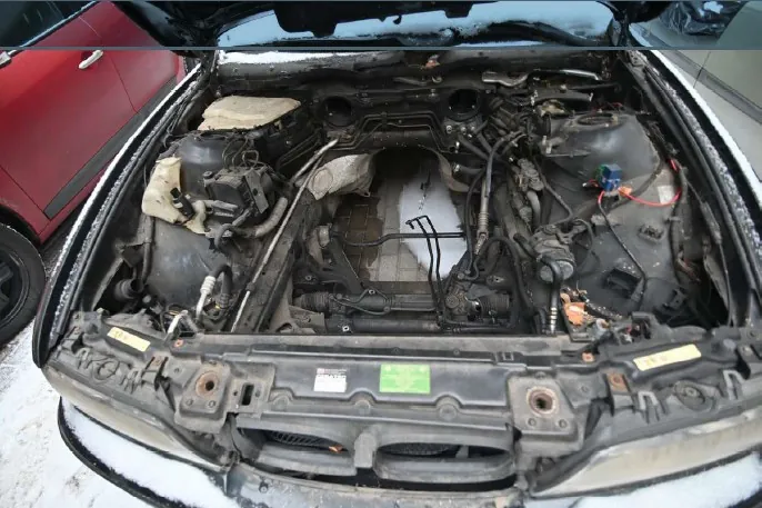 Biały samochód z otwartą maską ukazujący komponenty silnika, w tym przewody, zbiorniki i elementy mechaniczne. Na przedniej części komory silnika widoczna zielona naklejka oraz żółte etykiety. W tle fragment czerwonego samochodu. Śnieg pokrywa ziemię, krawędzie maski i części pojazdu.