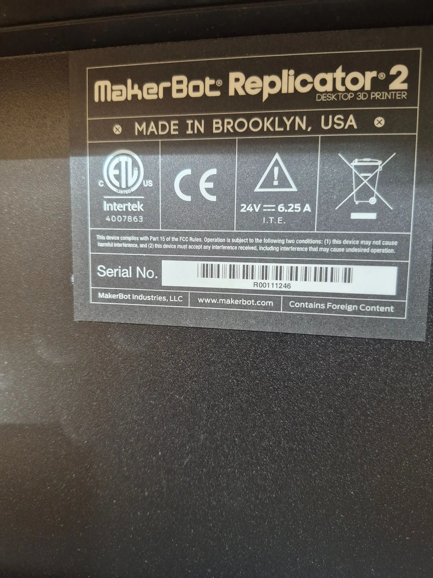 Zdjęcie srebrnej tabliczki znamionowej drukarki 3D MakerBot Replicator 2. Na górze znajduje się napis 'MakerBot Replicator 2 DESKTOP 3D PRINTER' oraz 'MADE IN BROOKLYN, USA'. Poniżej widoczne są certyfikaty i symbole: logo ETL Intertek z numerem 4007863, znak CE, trójkąt ostrzegawczy oraz symbol przekreślonego kosza na śmieci. Podana jest specyfikacja elektryczna: '24V ⎓ 6.25 A I.T.E.'. Poniżej znajduje się tekst dotyczący zgodności z przepisami FCC. Na dole widnieje napis 'Serial No.' obok kodu kreskowego i numeru seryjnego 'R00111246'. Na samym dole znajdują się informacje: 'MakerBot Industries, LLC', 'www.makerbot.com' oraz 'Contains Foreign Content'.