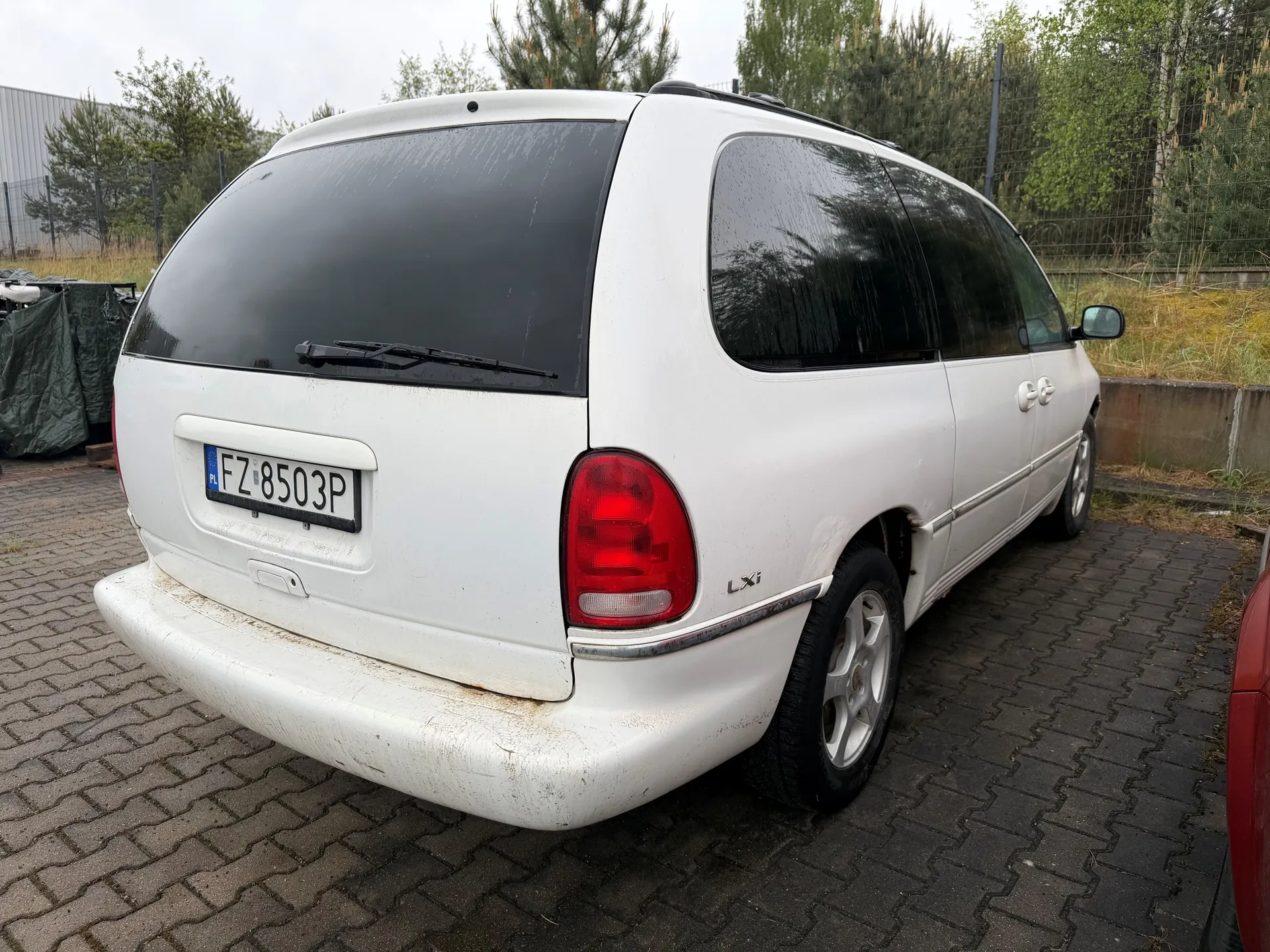 Tylny widok białego minivana z tablicą rejestracyjną FZ 8503P (PL) zaparkowanego na brukowanej powierzchni. Pojazd posiada ciemne szyby, czerwone światła tylne, wycieraczkę na tylnej szybie oraz napis 'LXi' na boku. W tle widoczne są drzewa, metalowe ogrodzenie, budynek przemysłowy oraz zielona płachta materiału po lewej stronie.