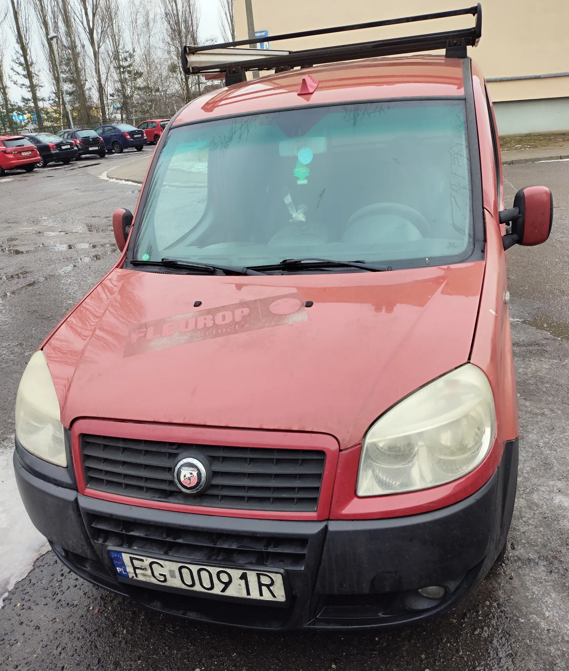 Czerwony samochód dostawczy z dachowym bagażnikiem, widok od przodu. Na maskie widoczny zatarty napis 'FLEUROP' oraz logo marki. Tablica rejestracyjna PL FG 0091R. W tle parking z innymi pojazdami, drzewami bez liści i budynkiem. Nawierzchnia asfaltowa z kałużami wody. W szybie przedniej wiszą zielone i niebieskie ozdoby.