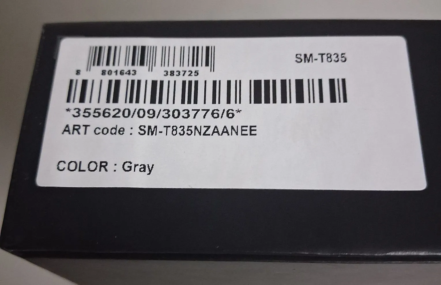 Zbliżenie na białą etykietę naklejoną na czarne pudełko. Na etykiecie znajdują się dwa kody kreskowe oraz teksty: 'SM-T835', '*355620/09/303776/6*', 'ART code : SM-T835NZAANEE' oraz 'COLOR : Gray'.
