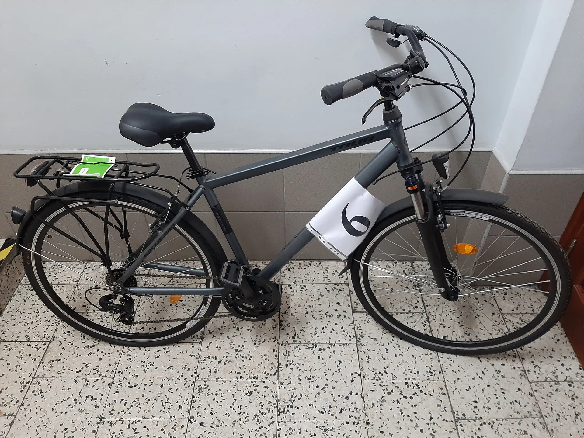 Szary rower typu trekkingowego z czarnym siedziskiem i kierownicą, stojący na podłodze z kafelkami w kropki. Na ramie widoczny napis 'TOUR' oraz biały pokrowiec z czarnym logo. Rower wyposażony w bagażnik tylny z zielonymi karteczkami, pomarańczowe odblaski na kołach i czarną oponę. Tło stanowi szara ściana z płytkami i fragment brązowej drzwi po prawej stronie.