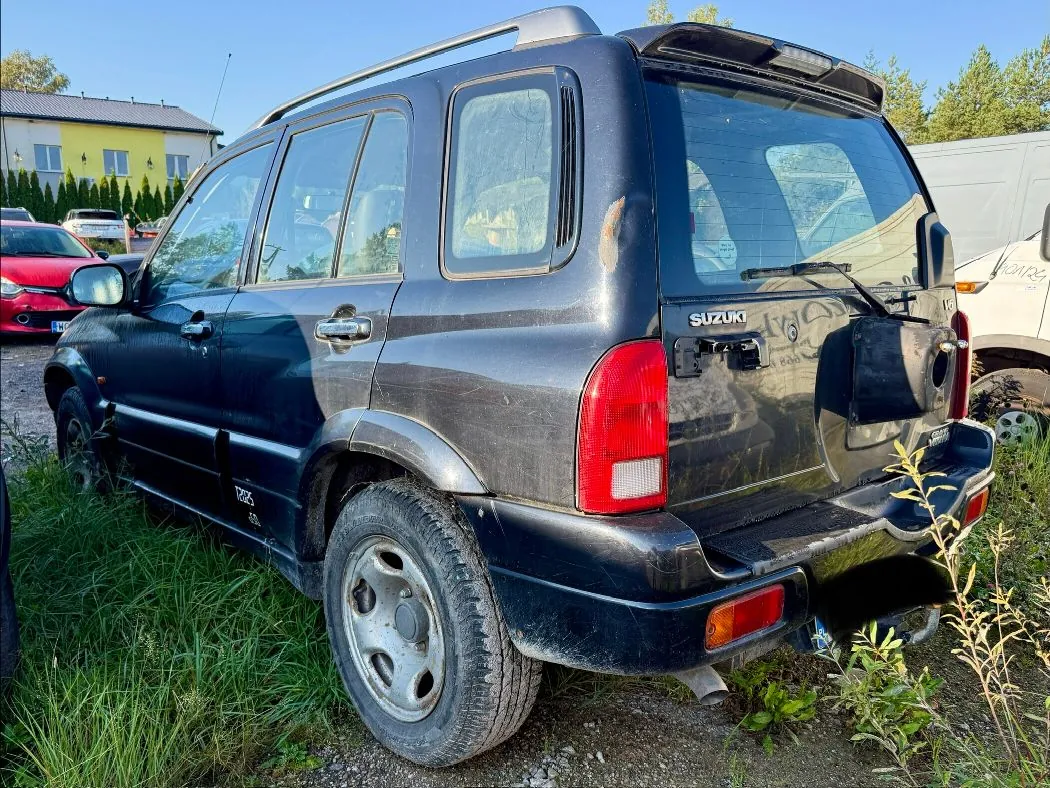 Czarny samochód terenowy marki Suzuki zaparkowany na trawiastym terenie. Pojazd jest uszkodzony, widoczne są wgniecenia na tylnym słupku oraz brak fragmentu dolnego progu po lewej stronie. Na tylnej klapie widnieje napis SUZUKI. W tle znajduje się żółty budynek oraz inny, czerwony samochód.
