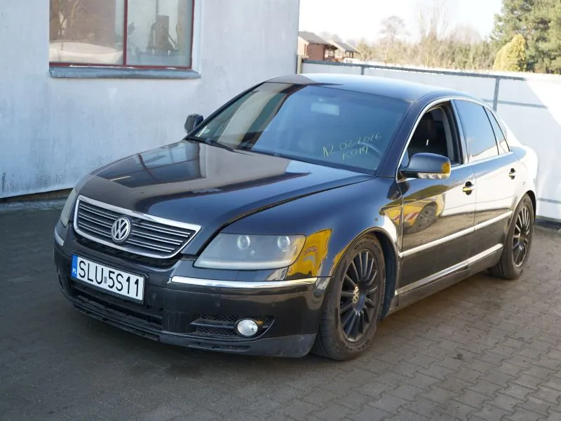 Czarny samochód osobowy marki Volkswagen zaparkowany na brukowanej powierzchni. Widoczna tablica rejestracyjna SLU 5S11 oraz napis na przedniej szybie: 12.02.2016 KOM. W tle budynek z oknem, ogrodzenie i drzewa. Samochód ma czarne felgi i reflektory.