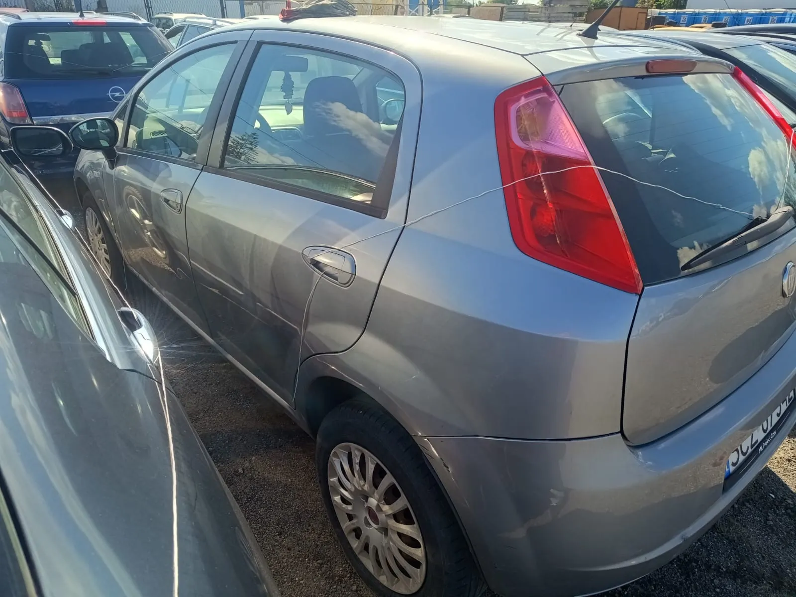 Srebrny samochód osobowy typu hatchback zaparkowany na placu obok innych pojazdów. Widok z tyłu i z boku auta, ukazujący tylną lampę, koło z felgą aluminiową oraz fragment tablicy rejestracyjnej.
