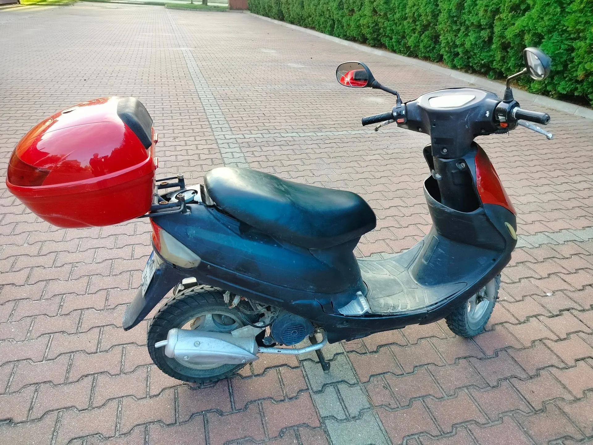 Czarny skuter zaparkowany na kostce brukowej. Pojazd posiada czerwony kufer zamontowany z tyłu oraz czarne siedzenie. W tle widoczny jest zielony żywopłot.