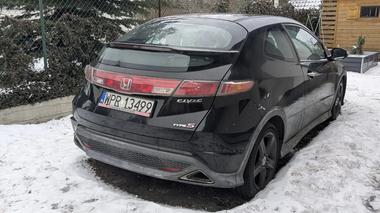 Czarny samochód Honda Civic Type S z tablicą rejestracyjną WPR 13499, zaparkowany na śnieżnej powierzchni. Widoczne są tylna część pojazdu, logo Hondy, napisy 'Civic' i 'Type S', czerwone światła oraz wydech. W tle siatka ogrodzeniowa, zasneżone krzewy, drewniana budowla i śnieg na ziemi.