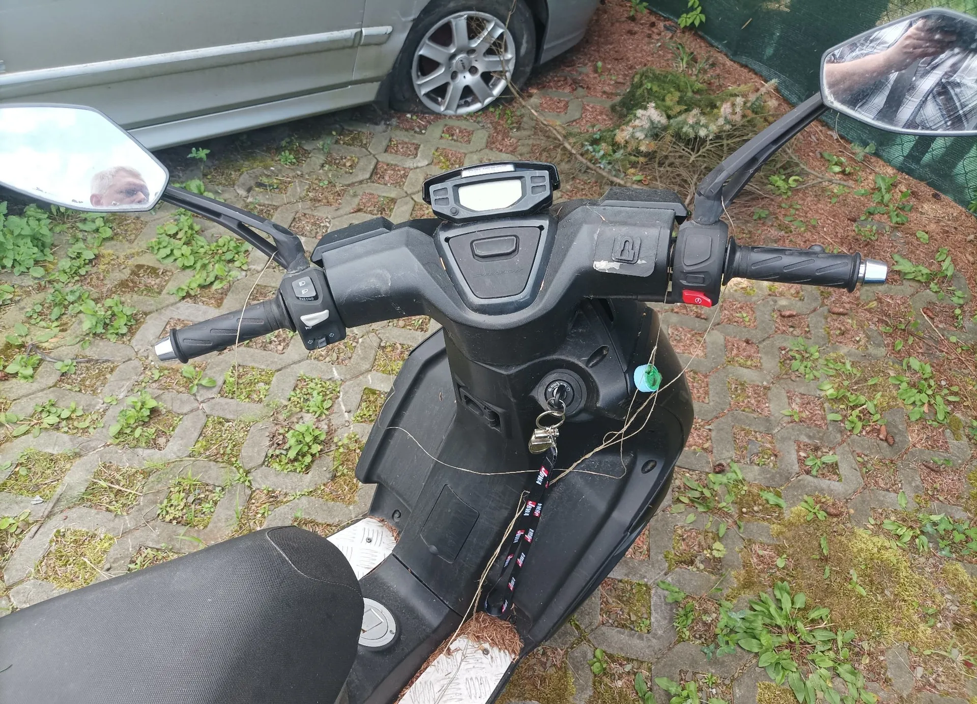 Widok z perspektywy kierowcy na czarny skuter zaparkowany na kostce brukowej z przerostami trawy. Widoczna jest kierownica z lusterkami, licznikiem, manetkami oraz stacyjką z włożonym kluczykiem i brelokiem. W tle widać fragment srebrnego samochodu.