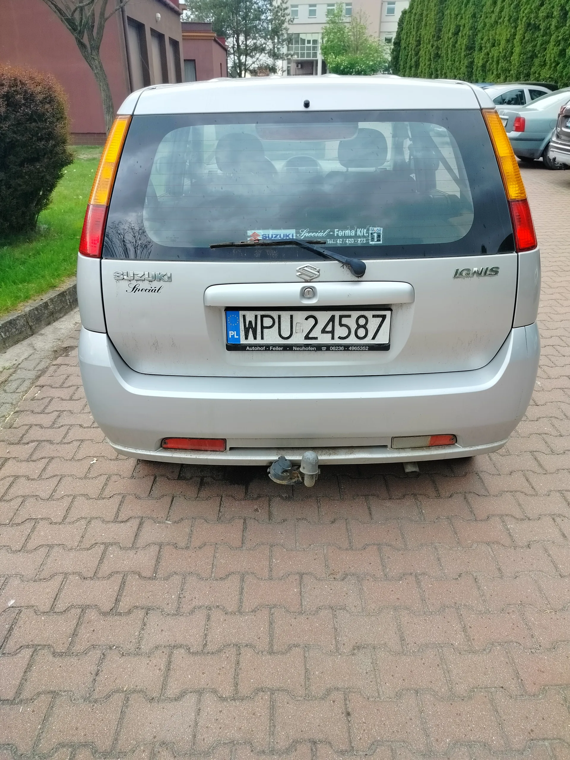Tył srebrnego samochodu Suzuki Ignis z tablicą rejestracyjną WPU 24587 (PL). Na szybie tylnej naklejka z napisem "Suzuki Special - Forma Kft." oraz numerami telefonów. Samochód zaparkowany na brukowanej powierzchni, w tle budynki, drzewa i inne pojazdy. Widoczne są również napisy "Suzuki Special" po lewej stronie i "Ignis" po prawej stronie nad klapą bagażnika.