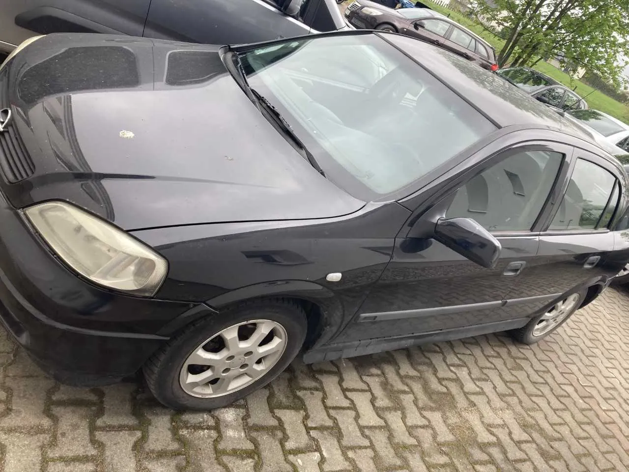 Czarny samochód osobowy typu sedan zaparkowany na brukowanej powierzchni. Widoczne są przednie światło, lusterko boczne oraz felgi. W tle znajdują się inne pojazdy, drzewa i trawnik. Na masce samochodu widoczne są drobne plamy.