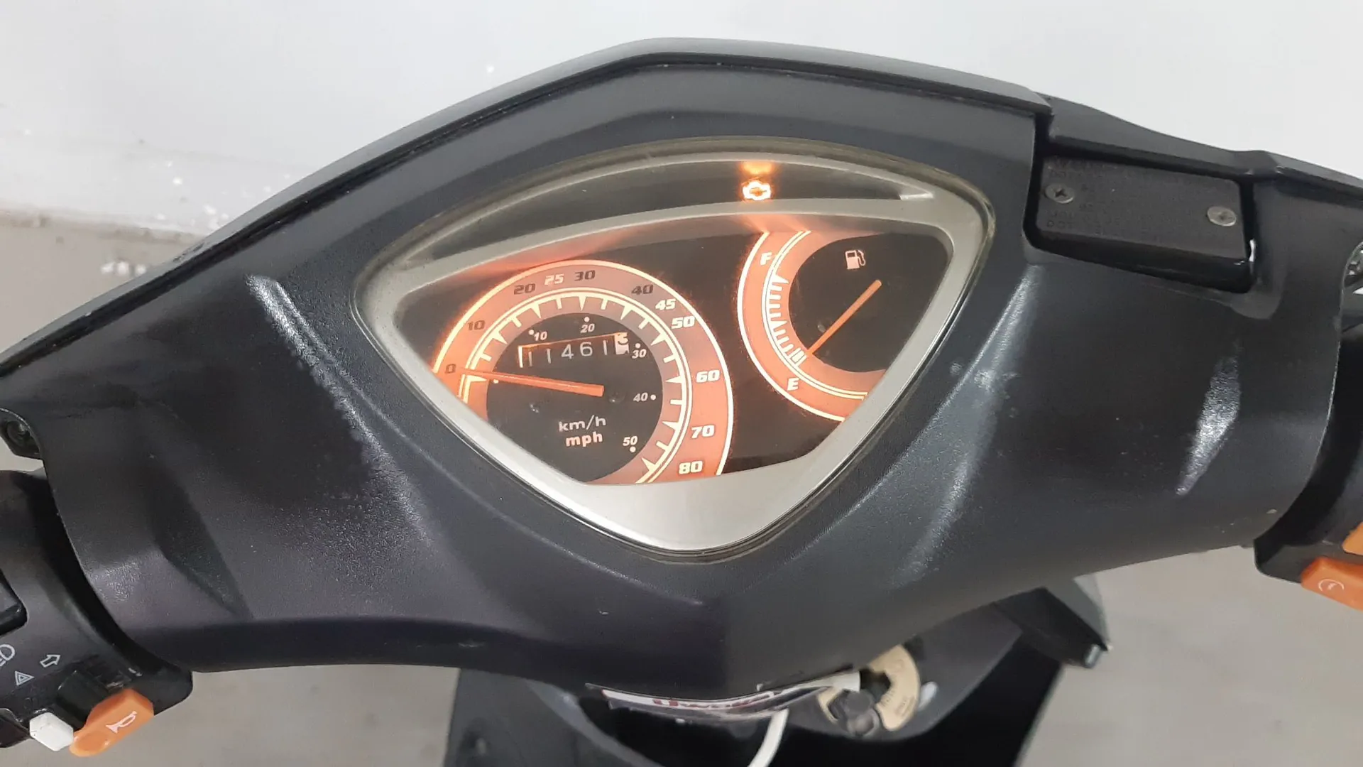 Zbliżenie na deskę rozdzielczą skutera. Widoczny jest analogowy prędkościomierz z pomarańczowym podświetleniem, skala sięga 80 km/h, a wskaźnik pokazuje wartość bliską 0. Wewnątrz prędkościomierza znajduje się licznik kilometrów z wartością 1461. Po prawej stronie znajduje się wskaźnik poziomu paliwa z oznaczeniami F (pełny) i E (pusty). Nad wskaźnikami świeci się pomarańczowa kontrolka w kształcie silnika.