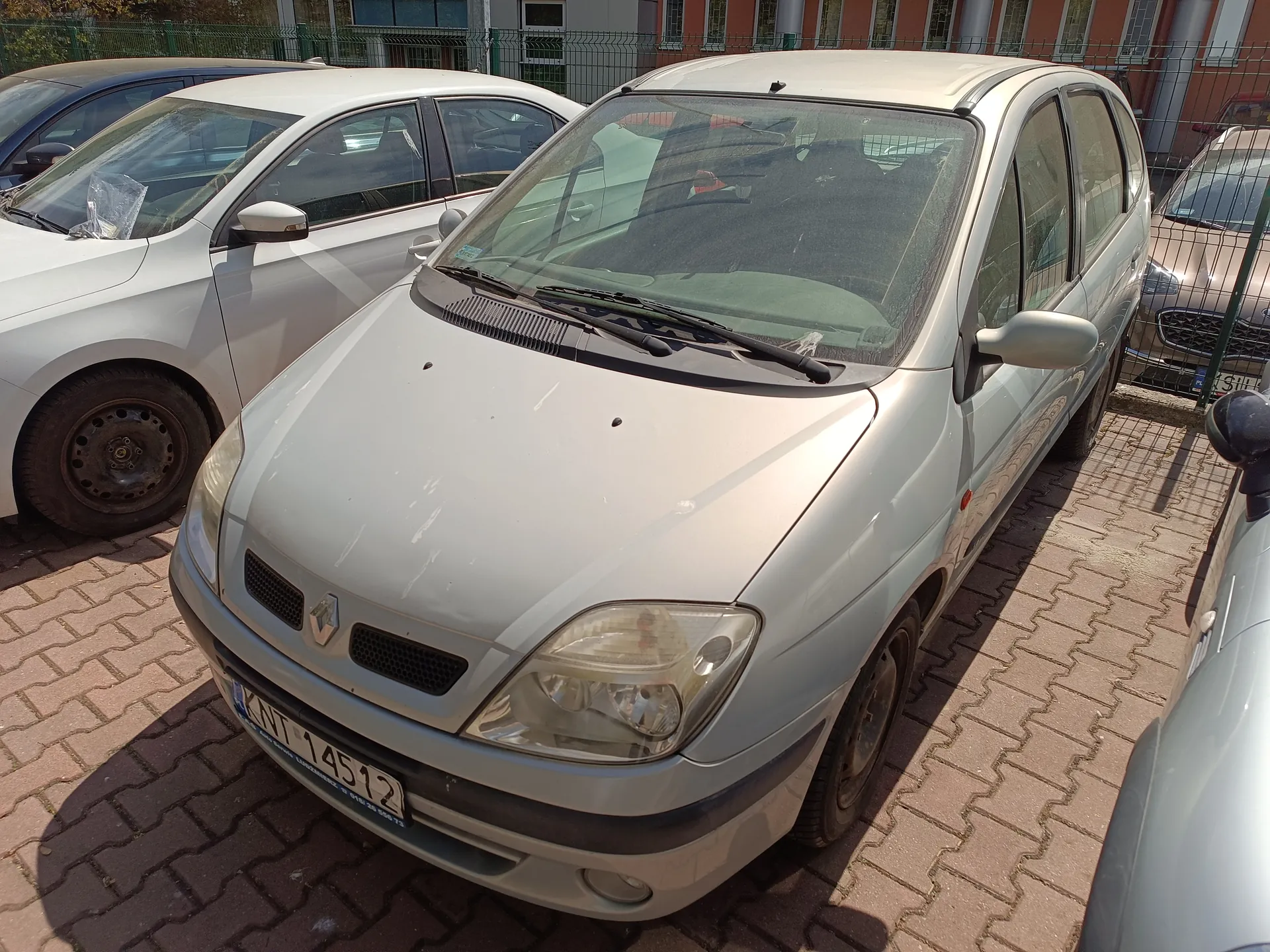 Srebrny samochód marki Renault z tablicą rejestracyjną KWT 14512 zaparkowany na brukowanej powierzchni. W tle inne pojazdy, ogrodzenie i ceglany budynek z oknami.