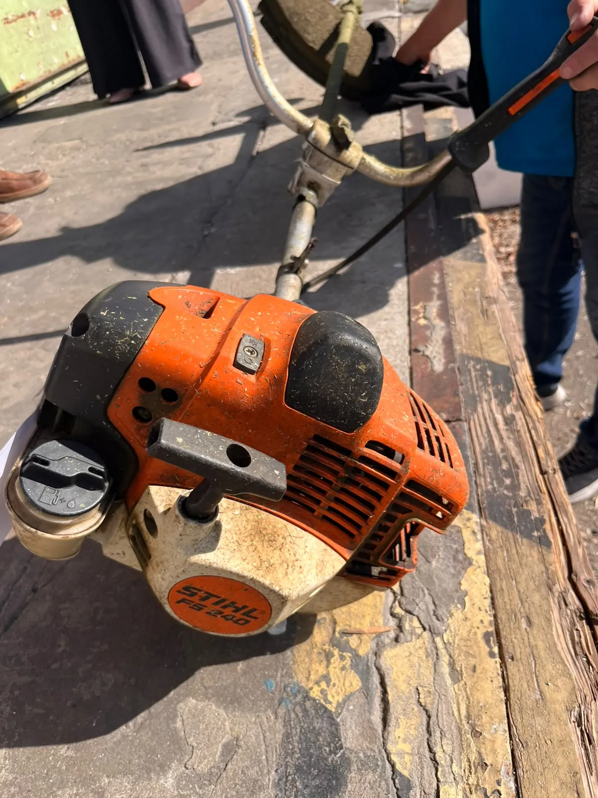 Pomarańczowa kosiarka spalinowa marki Stihl, model FS 240, leżąca na betonowym podłożu. Widoczny jest silnik z czarnymi elementami obudowy oraz długi drążek prowadzący do głowicy tnącej. W tle widoczne są nogi osób stojących obok urządzenia.