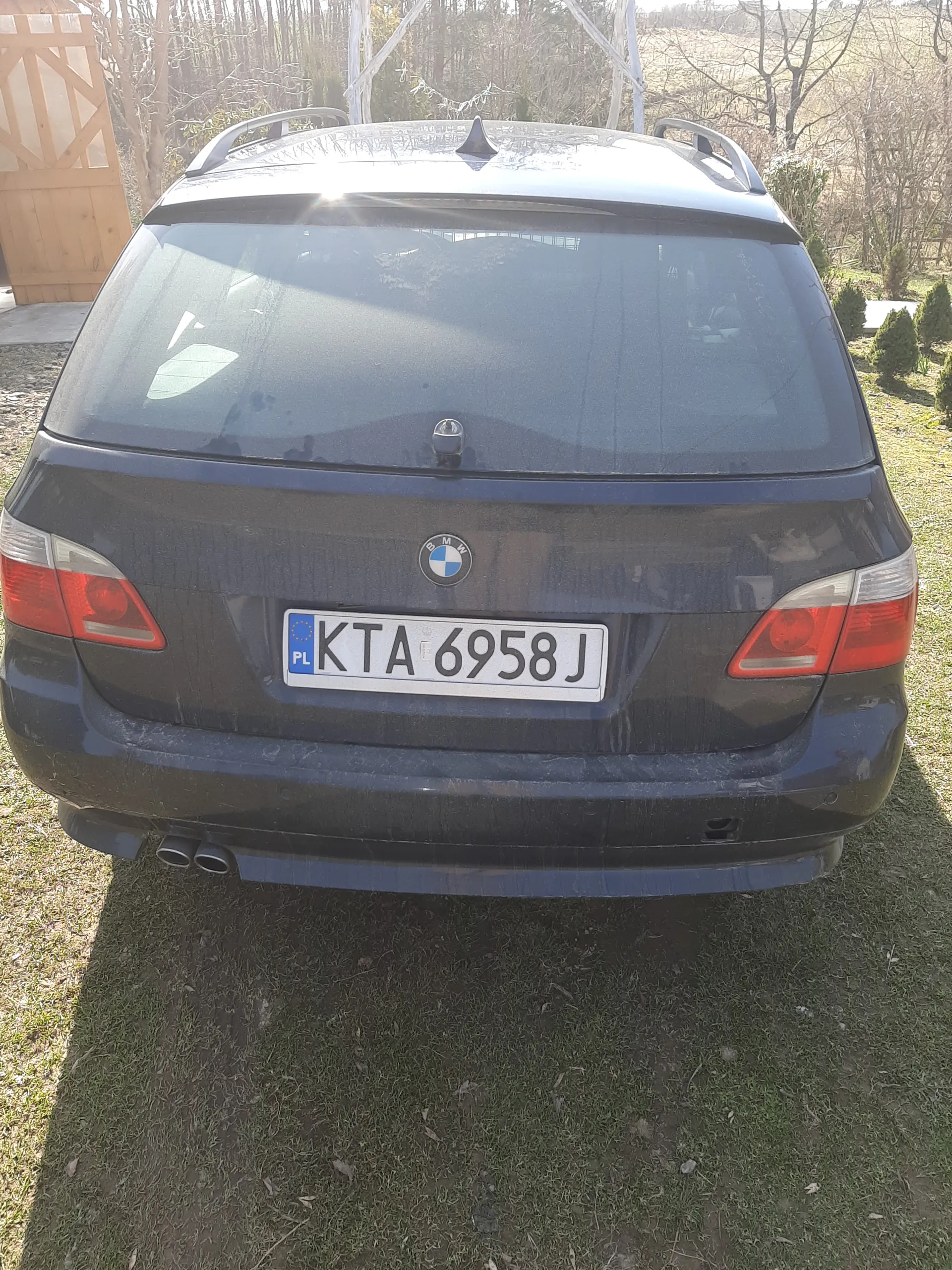 Tylny widok ciemnoniebieskiego samochodu marki BMW z tablicą rejestracyjną KTA 6958J (PL). Samochód zaparkowany na trawie, w tle widoczne drzewa, krzewy i drewniana brama.