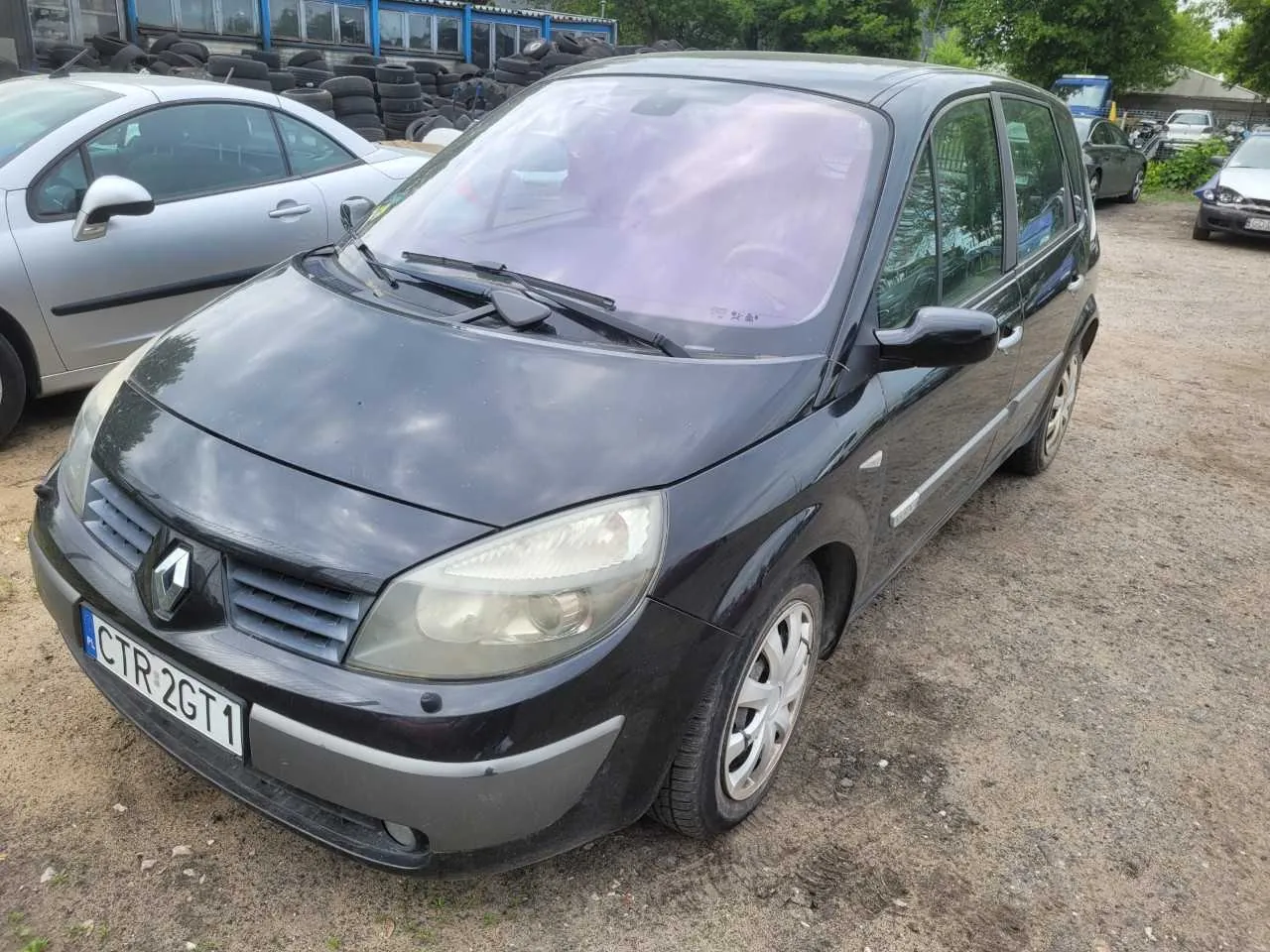 Czarny samochód marki Renault z tablicą rejestracyjną CTR 2GT1 zaparkowany na żwirowym placu. W tle srebrny samochód, stosy opon, budynki z niebieskimi oknami oraz drzewa.