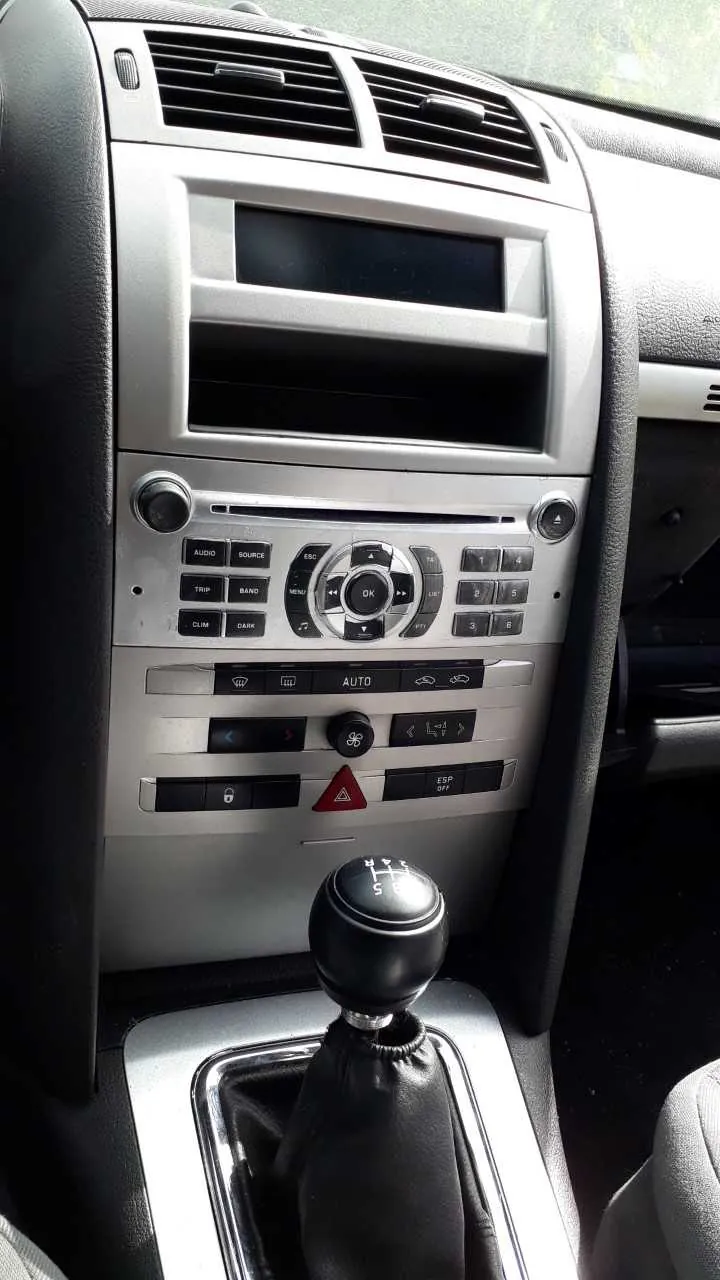 Wnętrze samochodu z widocznym panelem centralnym. Na górze znajdują się poziome wywiewacze powietrza, poniżej czarny ekran. Pod ekranem panel sterowania z przyciskami: AUDIO, SOURCE, TRIP, BAND, CLIM, DANK, przyciskiem centralnym OK oraz cyfrowymi przyciskami 1-6. Dalej widoczne są przyciski z napisami AUTO, ESP OFF, czerwony trójkąt ostrzegawczy oraz inne symbole. Na dole znajduje się czarna dźwignia zmiany biegów z oznaczeniami R, 1, 2, 3, 4, 5. Całość wykonana w kolorze srebrnym i czarnym.