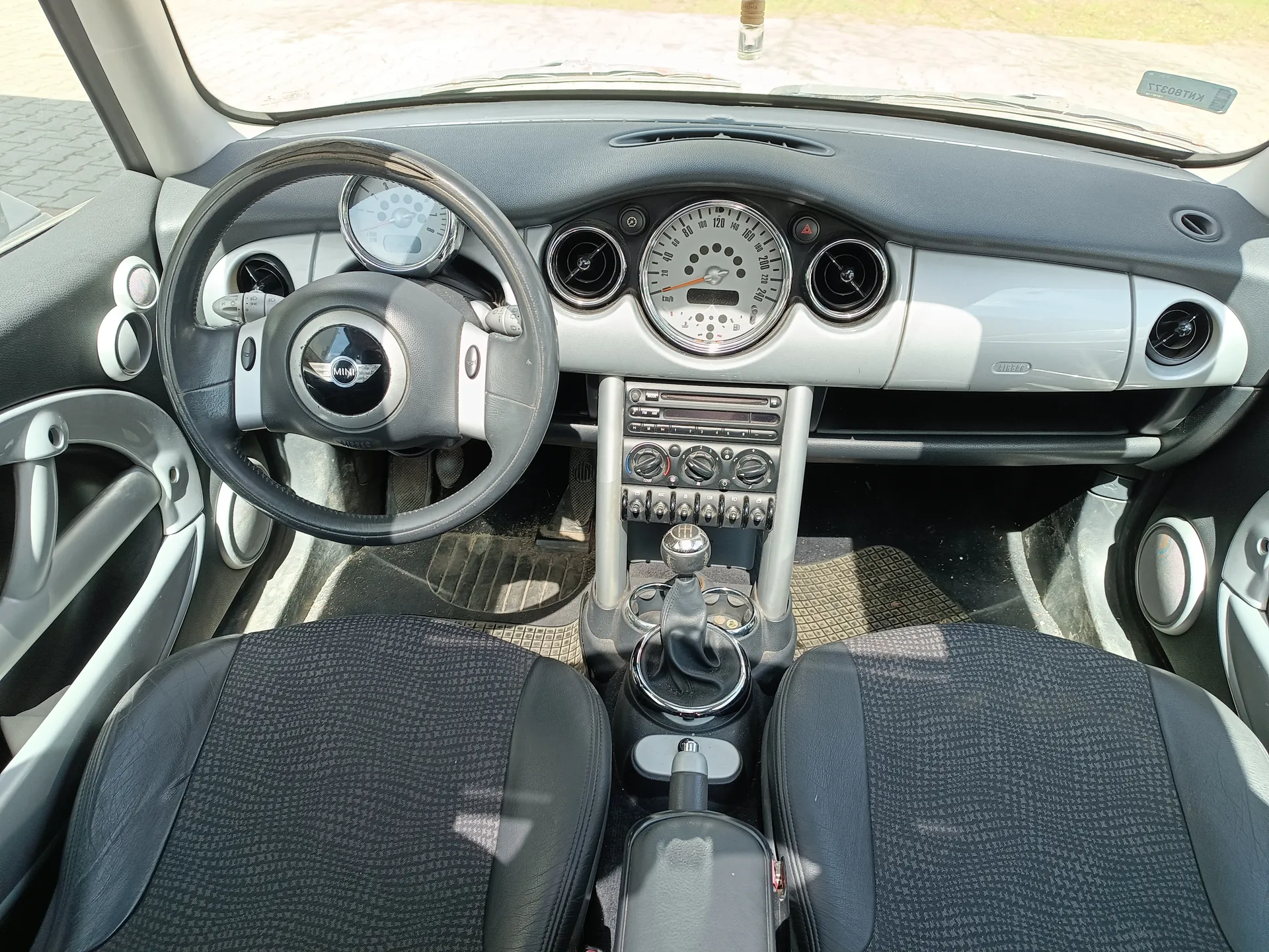 Wnętrze samochodu marki MINI z widokiem na kierownicę z logo MINI, czarno-srebrną deską rozdzielczą z okrągłymi wskaźnikami, w tym prędkościomierzem, centralną konsolą z przyciskami i pokrętłami, dźwignią zmiany biegów oraz czarnymi siedzeniami z szarym wzorem. Widoczne są również drzwi z uchwytem i głośnikiem, a przez przednią szybę widać fragment zewnętrznej nawierzchni.