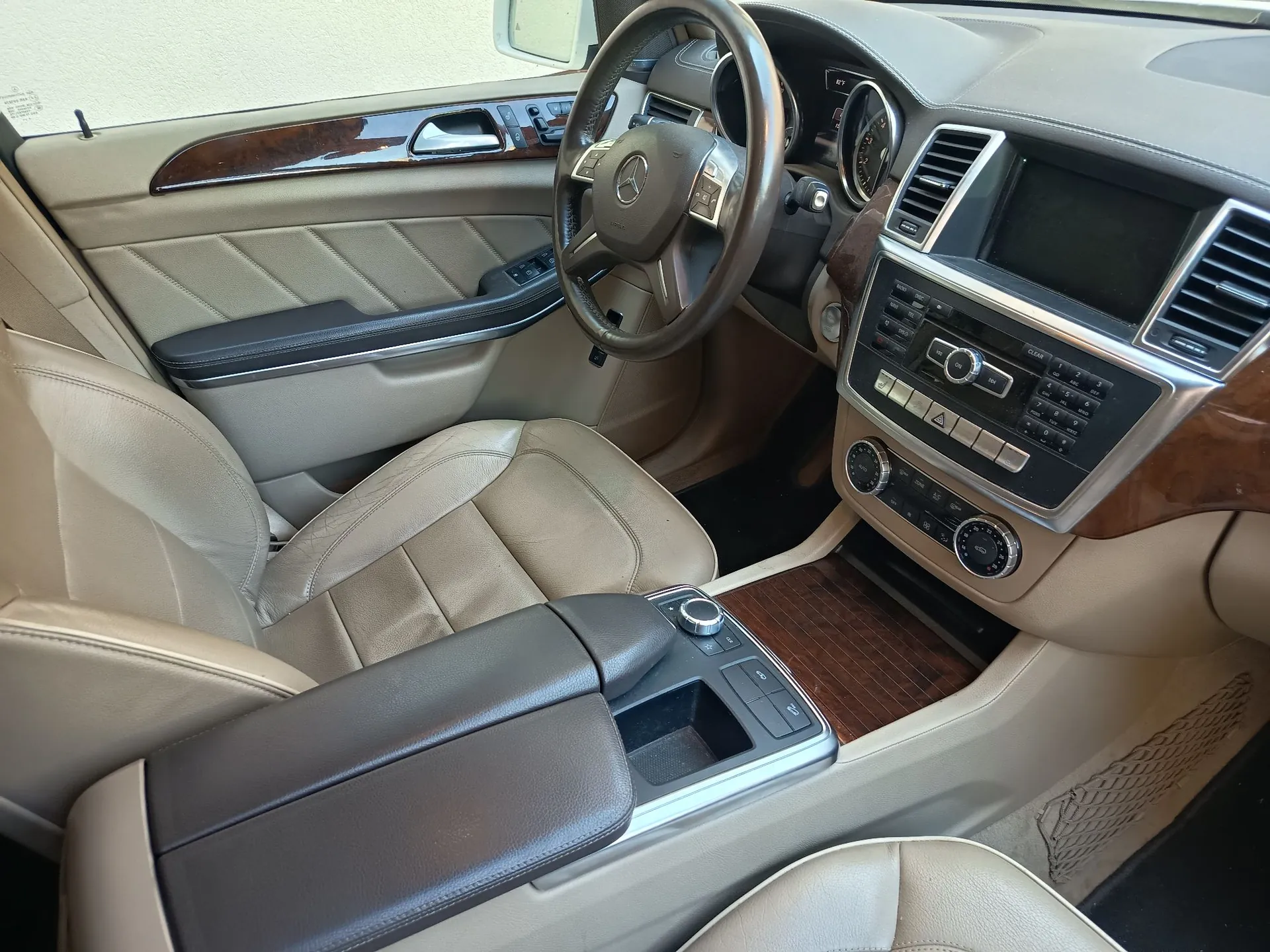 Wnętrze samochodu marki Mercedes-Benz z perspektywy tylnego siedzenia. Widoczny jest przedni rząd siedzeń w kolorze beżowym, ciemnobrązowa skórzana kierownica z logo Mercedes na środku, deska rozdzielcza z ekranem multimedialnym, przyciskami sterującymi i drewnianymi wykończeniami. Pomiędzy przednimi fotelami znajduje się ciemnobrązowy podłokietnik oraz konsola środkowa z drewnianym panelem.