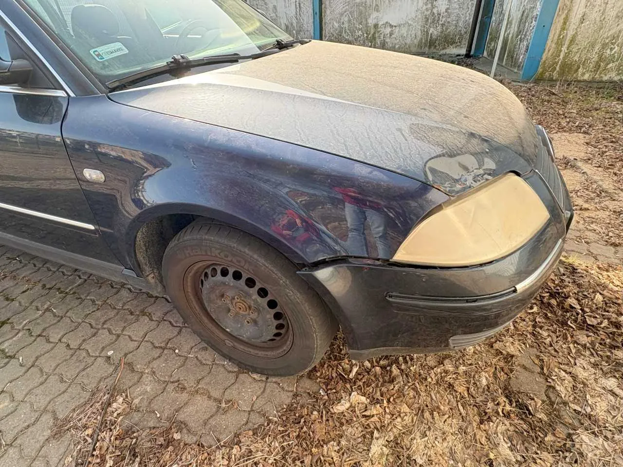 Ciemnoniebieski samochód osobowy z widocznym uszkodzeniem przedniego zderzaka i lewego reflektora, który jest częściowo zakryty beżową taśmą. Na przednim lewym kole widoczna brudna opona z rdzewiejącym felgiem. Samochód zaparkowany na brukowanej powierzchni, wokół sucha trawa i liście. W tle zniszczona ściana z niebieskimi elementami konstrukcyjnymi. Na przedniej szybie naklejka z nieczytelnym tekstem.