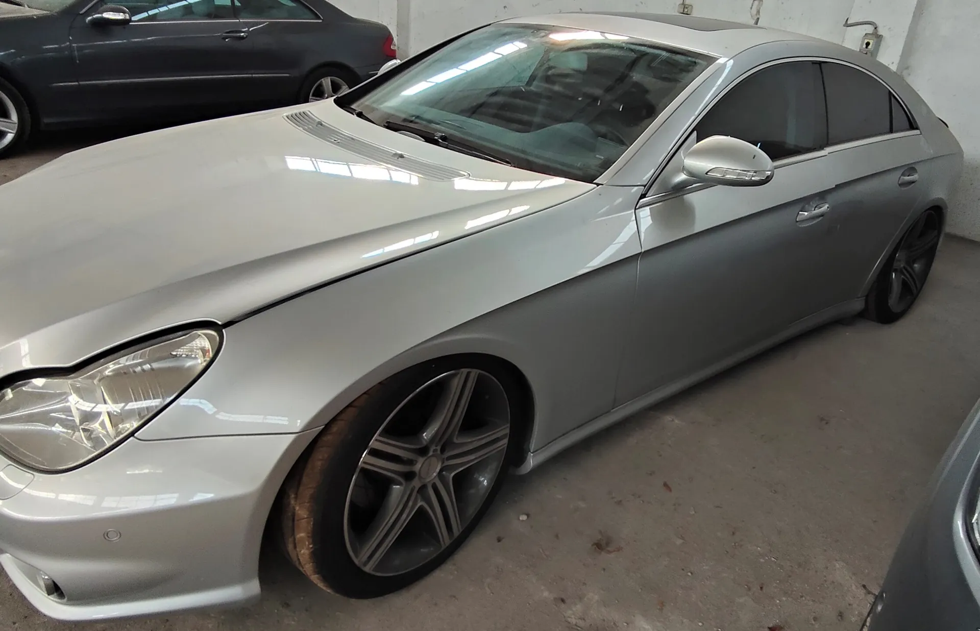 Srebrny samochód osobowy typu coupe zaparkowany w garażu. Widok z przodu i z boku, ukazujący maskę, przedni reflektor, felgi aluminiowe z pięcioma ramionami oraz linię nadwozia. W tle widoczny jest fragment innego, ciemnego samochodu.