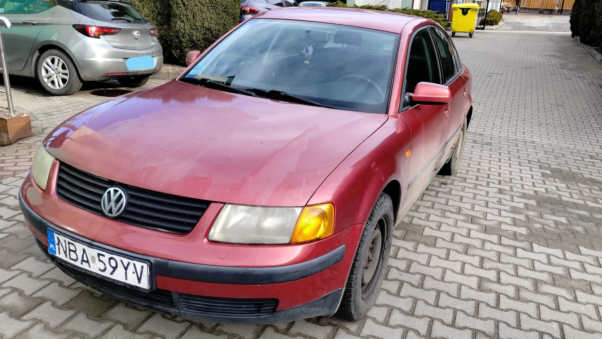 Czerwony samochód marki Volkswagen z tablicą rejestracyjną NBA 59YV z flagą PL. Samochód stoi na brukowanej powierzchni. W tle widoczny srebrny samochód, żółty pojemnik na śmieci, krzewy oraz budynki. Tablica rejestracyjna i tekst na niej są czytelne i ustawione poziomo.