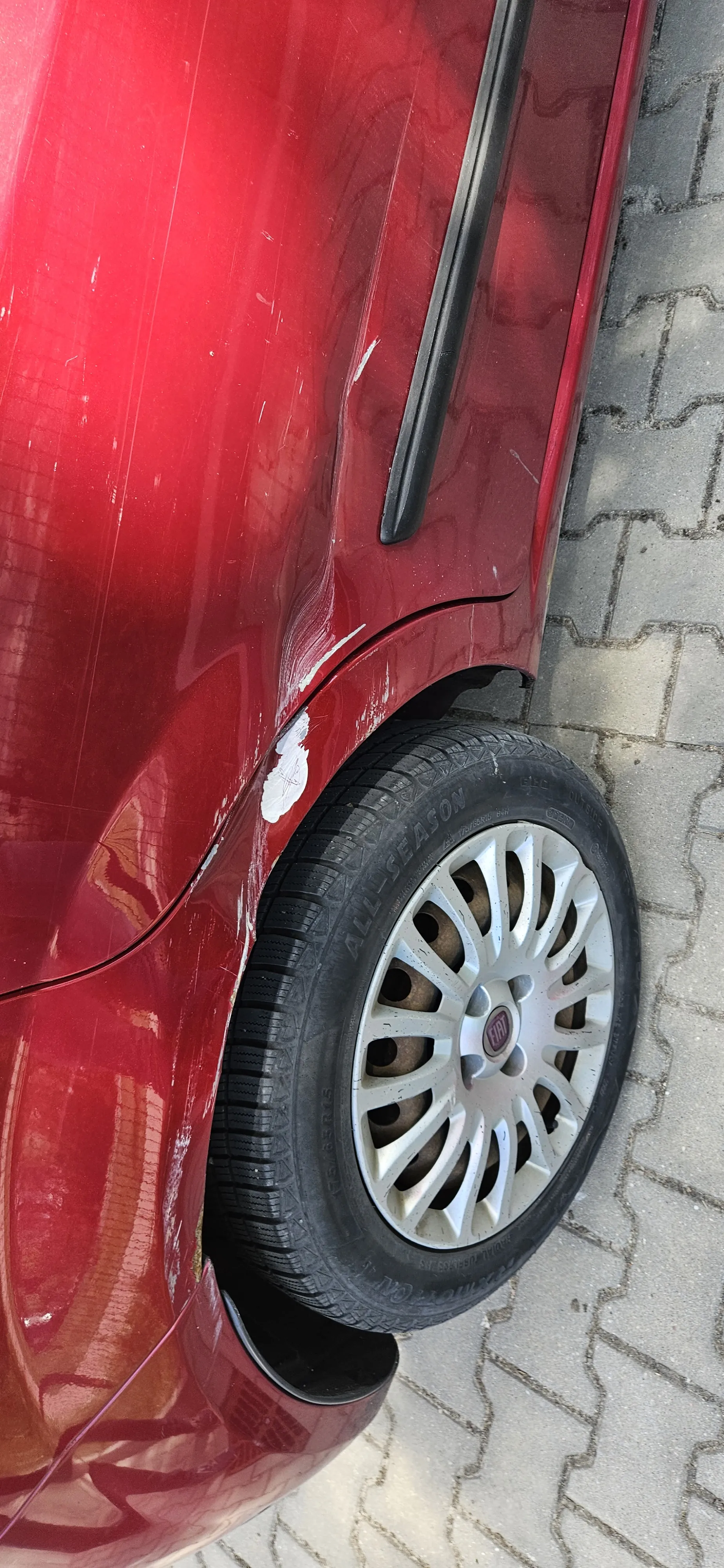 Czerwony samochód z widocznymi zadrapaniami i białymi plamami farby na tylnej części. W tylnej części umieszczono oponę z srebrnym felgiem z logo w centrum. Tło stanowi brukowana nawierzchnia z kamiennymi płytami.
