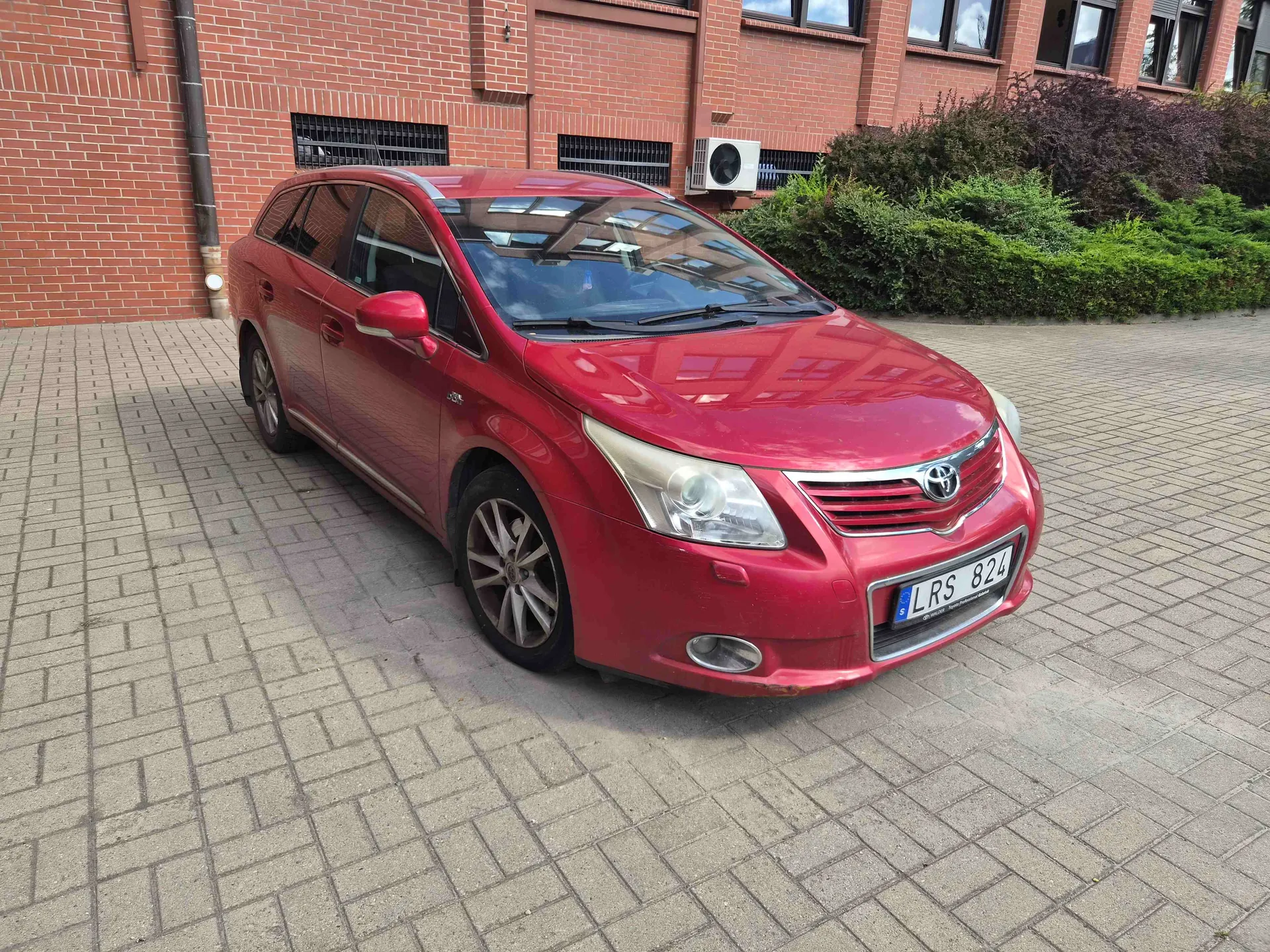 Czerwony samochód marki Toyota w nadwoziu kombi zaparkowany na kostce brukowej przed budynkiem z czerwonej cegły. Samochód ma polską tablicę rejestracyjną o numerze LRS 824. W tle widoczne są zielone krzewy oraz jednostka zewnętrzna klimatyzatora zamontowana na ścianie budynku.