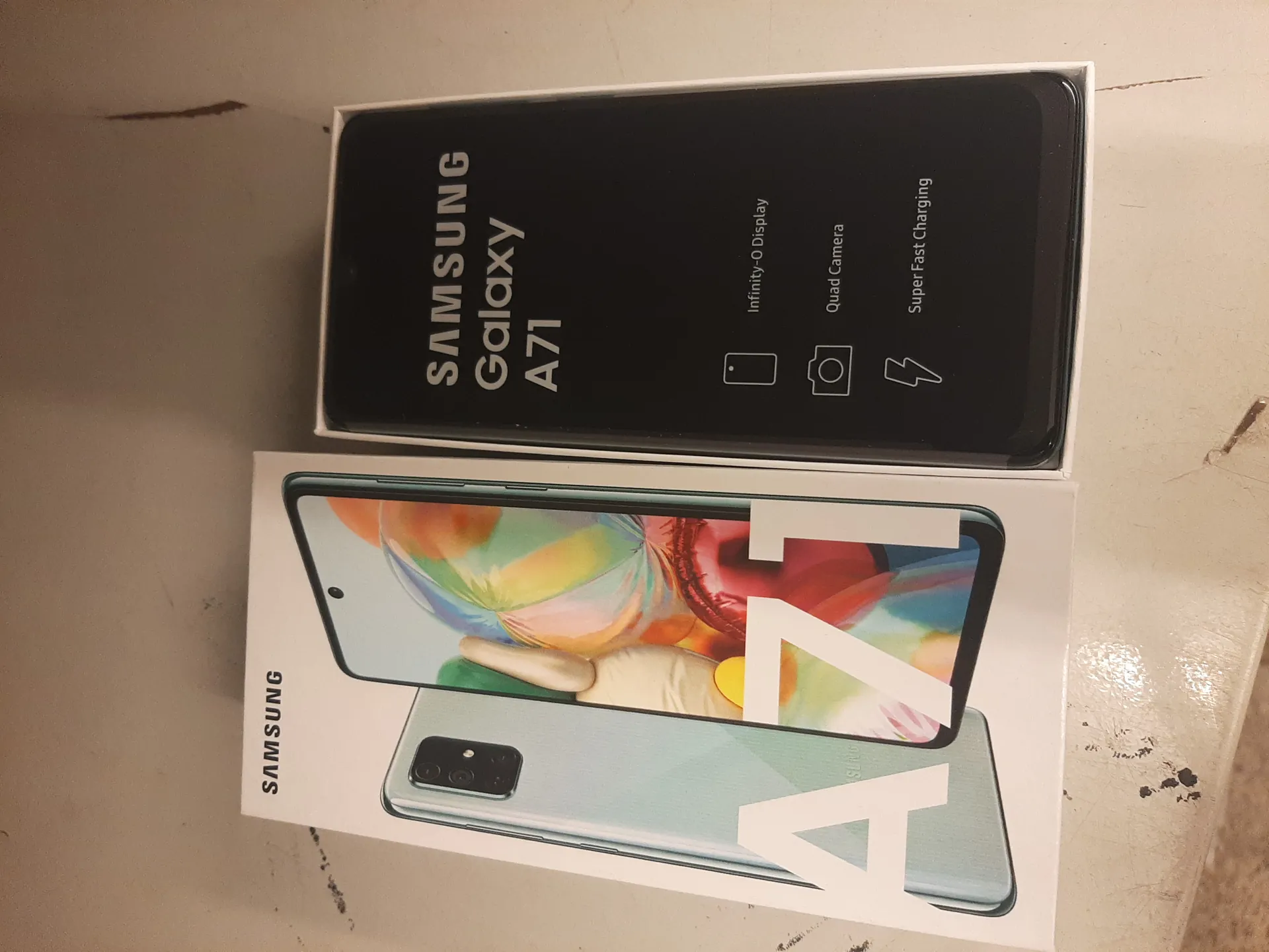 Na zdjęciu znajdują się dwa pudełka produktowe Samsung Galaxy A71. Po lewej stronie białe pudełko z obrazem telefonu w kolorze jasnoniebieskim, przedstawiającym przód i tył urządzenia. Na pudełku widnieje napis 'SAMSUNG' oraz duży napis 'A71'. Po prawej stronie czarne pudełko z białym tekstem: 'SAMSUNG Galaxy A71', poniżej ikony i opisy funkcji: 'Infinity-O Display', 'Quad Camera', 'Super Fast Charging'. Tło stanowi jasna powierzchnia z widocznymi plamami i zadrapaniami.