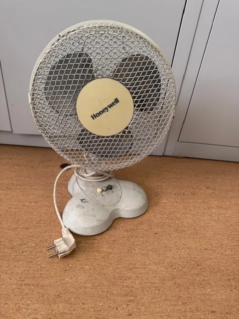 Biały, plastikowy wentylator marki Honeywell z okrągłą osłoną z metalowej siatki. Wentylator stoi na brązowej powierzchni, a obok niego leży biały kabel z wtyczką elektryczną. Na środku osłony znajduje się żółtawa tarcza z napisem 'Honeywell'. Podstawa wentylatora posiada dwa przyciski sterujące.