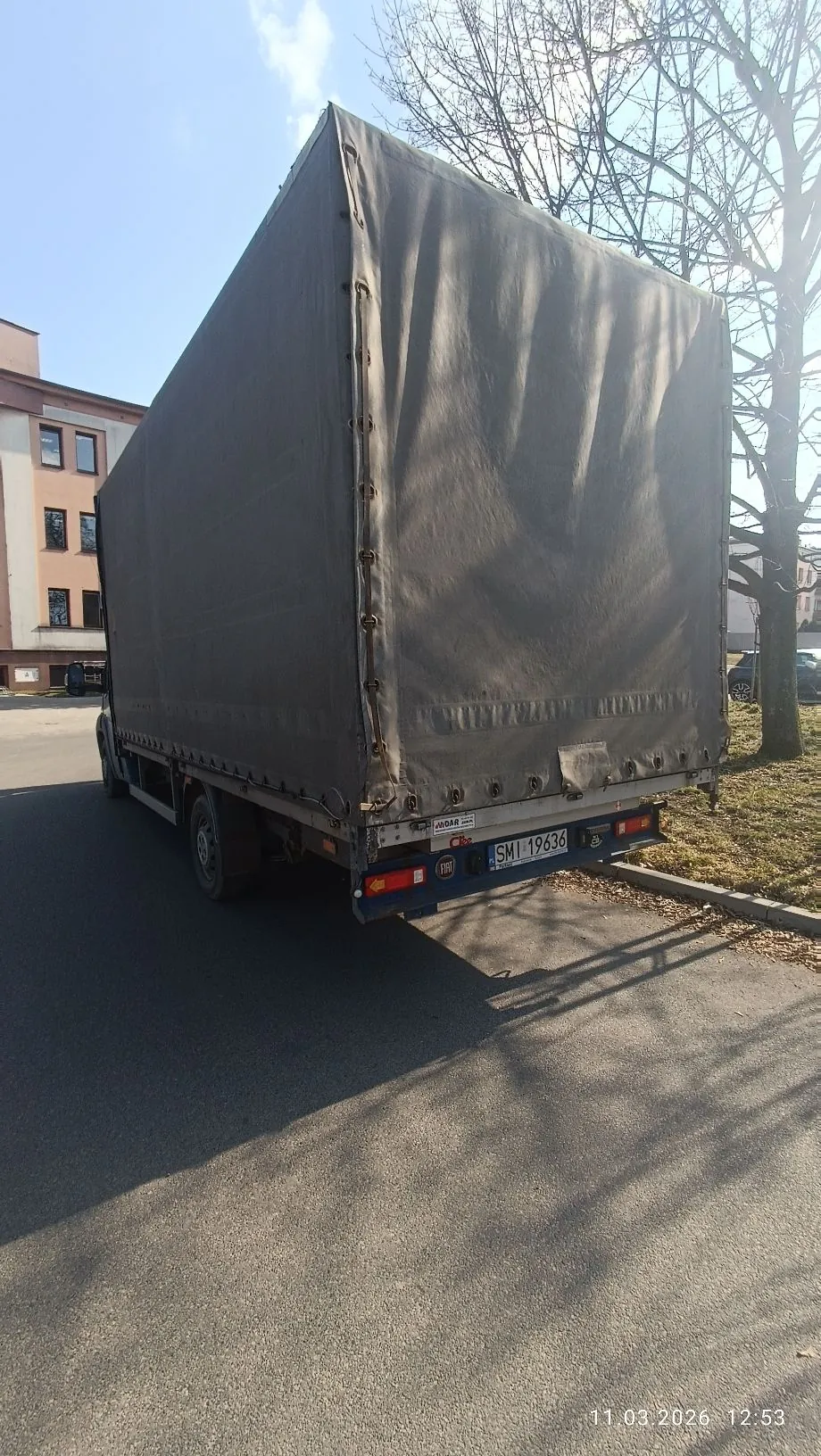 Zdjęcie tyłu białej ciężarówki z szarym plandekowym nadwoziem, zaparkowanej na asfaltowej drodze. Pojazd posiada polską tablicę rejestracyjną o numerze SMI 19636. W tle widoczny jest jasny budynek oraz bezlistne drzewo. W prawym dolnym rogu znajduje się data i godzina: 11.03.2026 12:53.
