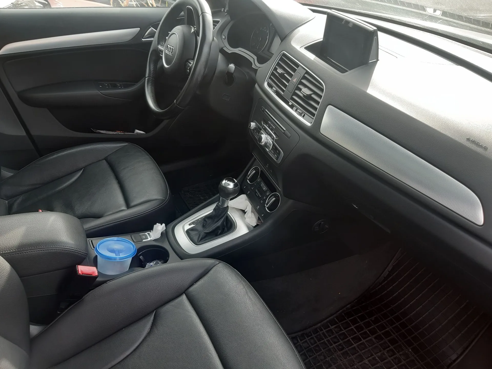 Wnętrze samochodu z widokiem na kokpit, czarną skórzaną tapicerkę i deskę rozdzielczą. Widoczna jest kierownica z logo Audi, lewarek zmiany biegów, dwa przednie fotele oraz konsola środkowa, w której znajduje się niebieski plastikowy pojemnik. Na górze deski rozdzielczej umieszczony jest uchwyt na telefon.