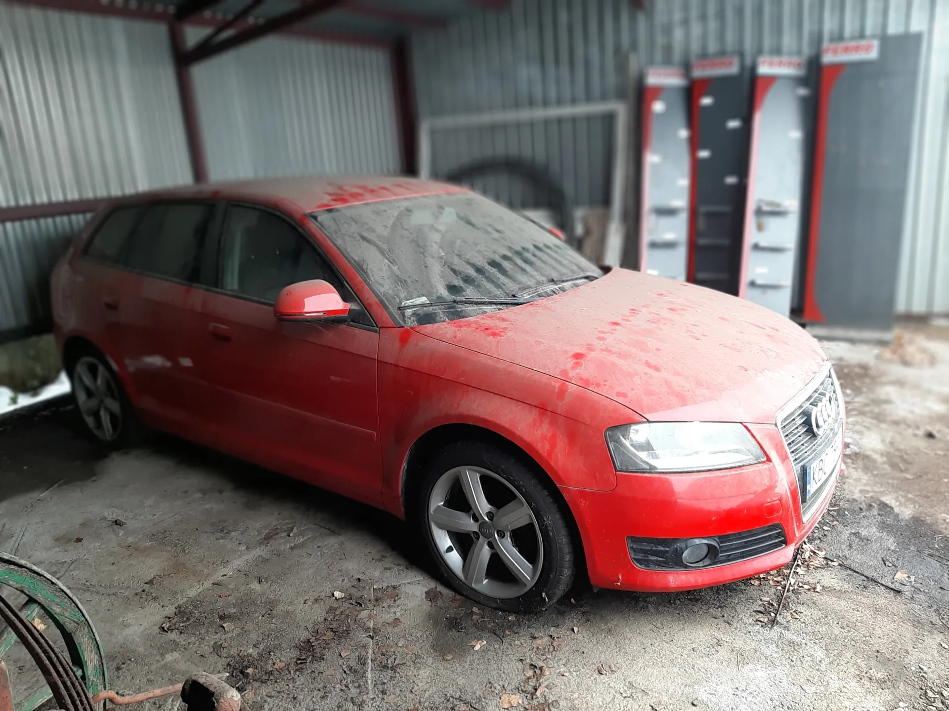 Czerwony samochód Audi kombi, pokryty brudem i czerwonymi plamami, zaparkowany na betonowej powierzchni w przemyślnym pomieszczeniu z metalowymi ścianami. W tle widoczne są pionowe konstrukcje z czerwonymi elementami oraz fragment zielonej opony w lewym dolnym rogu. Samochód posiada czyste reflektory, srebrne felgi i zanieczyszczoną przednią szybę.