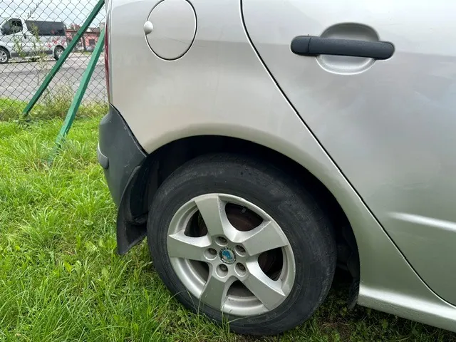 Zbliżenie na tylne prawe koło srebrnego samochodu zaparkowanego na trawie obok zielonego ogrodzenia z siatki. Widoczny jest fragment nadwozia z klamką drzwi i klapką wlewu paliwa. W tle widać inny, biały samochód.
