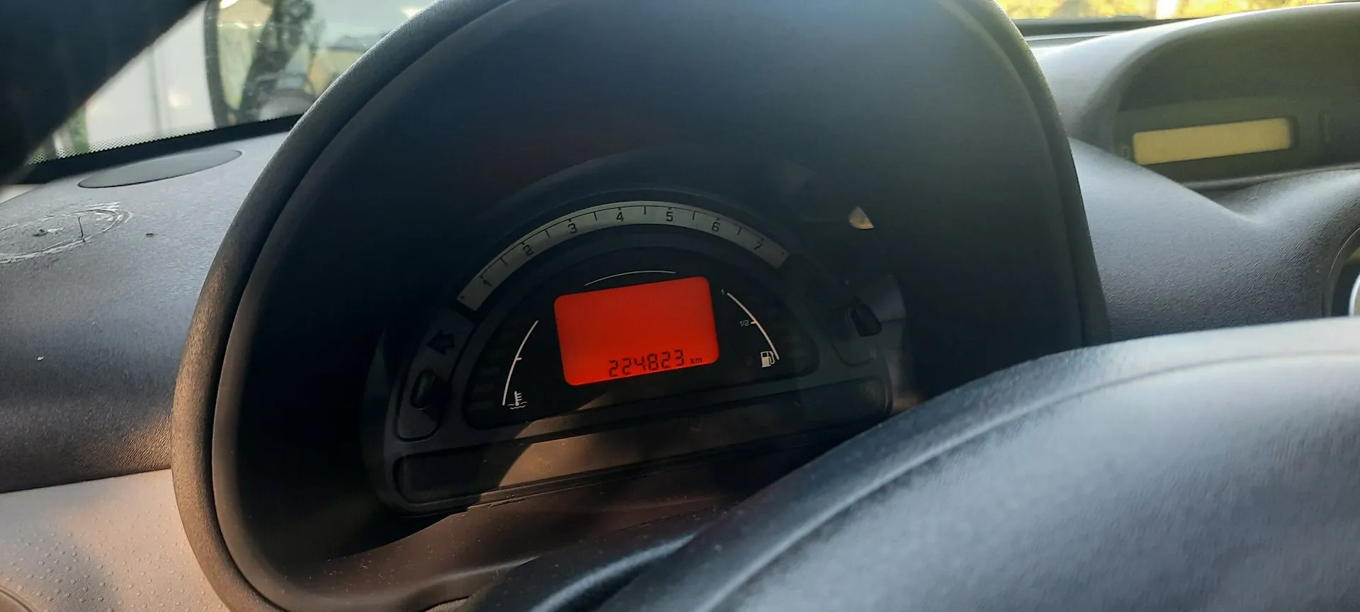 Zbliżenie na deskę rozdzielczą samochodu z widocznym licznikiem kilometrów. Na czerwonym, cyfrowym wyświetlaczu widnieje liczba 224823 km. Wokół wyświetlacza znajdują się wskaźniki paliwa oraz obrotomierza z cyframi od 1 do 7.