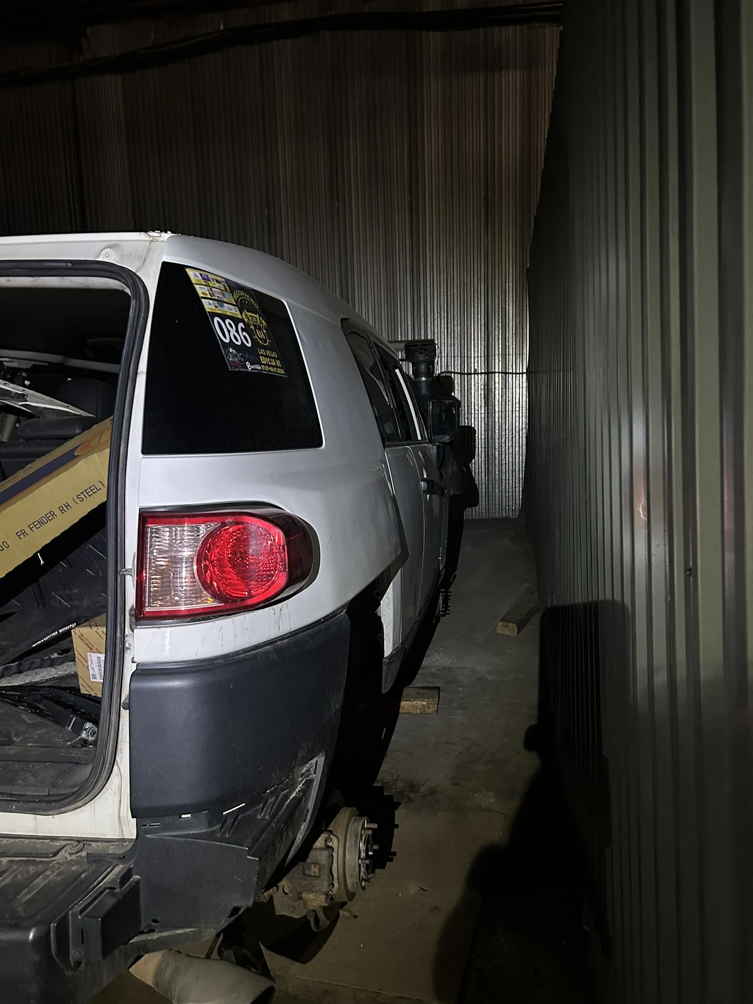 Biały SUV zaparkowany w ciemnym garażu z metalowymi ścianami. Tylna klapa pojazdu jest otwarta, widoczne są pudełka, w tym jedno z napisem 'FR FENDER RH (STEEL)'. Na tylnej szybie naklejka z numerem '086' oraz dodatkowym tekstem. Samochód nie ma jednego tylnego koła, odsłaniając mechanizm hamulcowy. Tylne światła są czerwone, a podwozie częściowo uszkodzone. Podłoga garażu jest betonowa, a w tle widoczne są pionowe blachy metalowe.