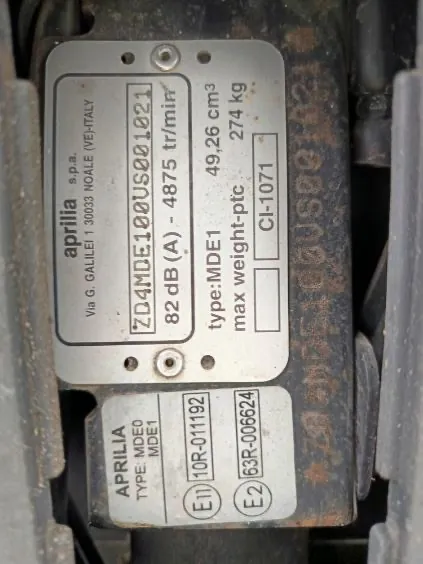 Dwa metalowe tablice z napisami zamocowane na powierzchni. Górną tablicę zajmuje napis 'aprilialia s.p.a. Via G. GALILEI 1 30033 NOALE (VE) ITALY', poniżej kod 'ZD4MDE100US001021', '82 dB (A) - 4875 tr/min', '49,26 cm³', '274 kg', 'type:MDE1', 'max weight-ptc' oraz 'CI-1071'. Dolna tablica zawiera 'APRILIA', 'TYPE MDE0', 'MDE1', oznaczenia 'E1 10R-011192' i 'E2 63R-006624'. Tekst na obu tablicach jest poziomy i czytelny.