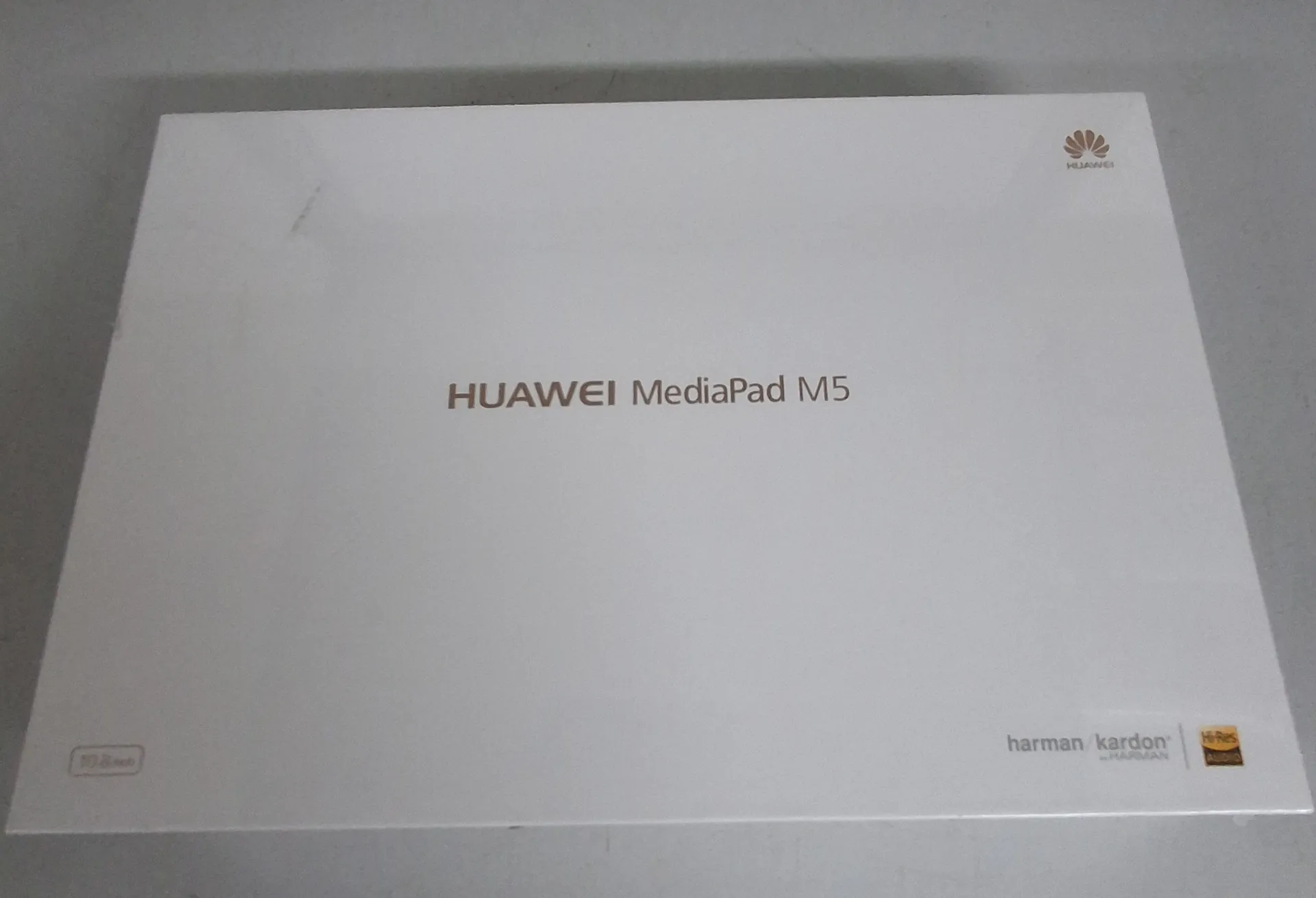 Białe, prostokątne pudełko z tabletem. Na środku znajduje się napis 'HUAWEI MediaPad M5'. W prawym górnym rogu widoczne jest logo Huawei. W prawym dolnym rogu znajdują się logotypy 'harman/kardon' oraz 'Hi-Res Audio'. W lewym dolnym rogu widnieje napis '10.8 inch'.