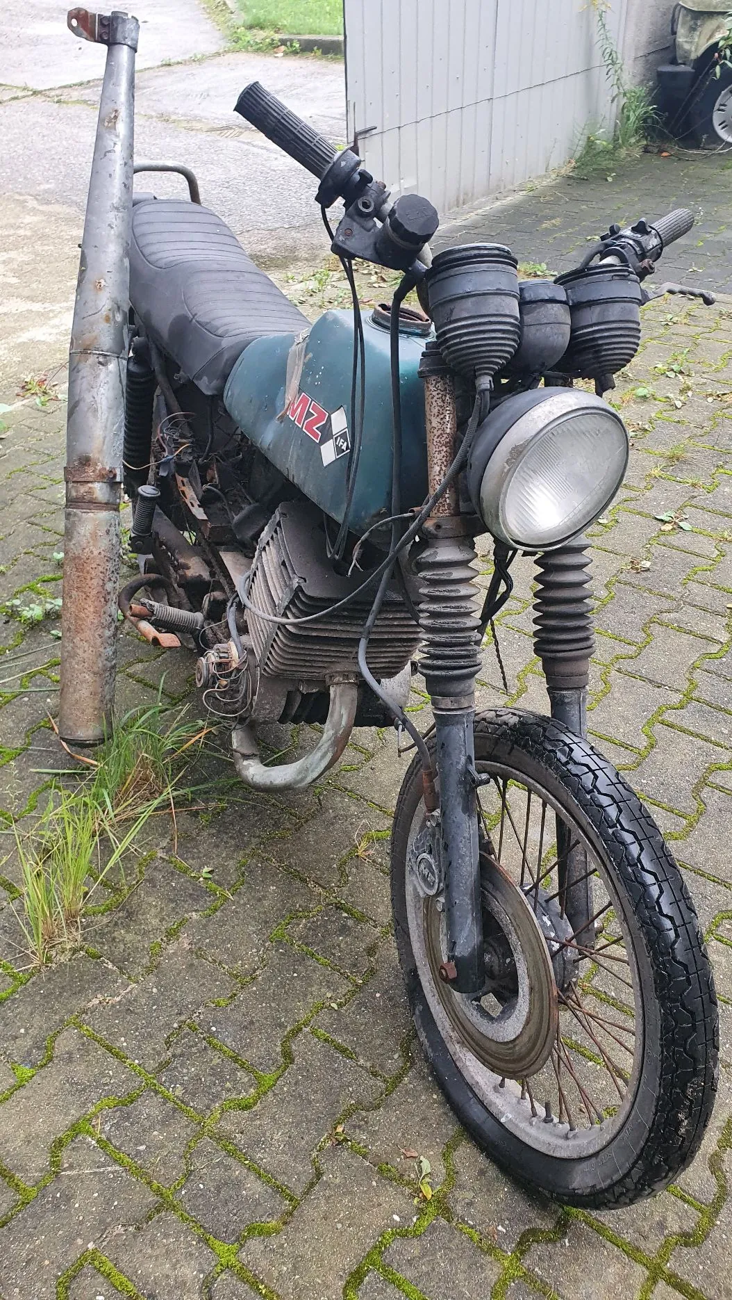 Stary, zniszczony motocykl marki MZ w kolorze ciemnozielonym, zaparkowany na kostce brukowej porośniętej mchem. Pojazd jest w złym stanie technicznym, z widoczną rdzą na rurze wydechowej i elementach ramy. Widoczny jest okrągły reflektor, kierownica z manetkami oraz charakterystyczny silnik dwusuwowy.