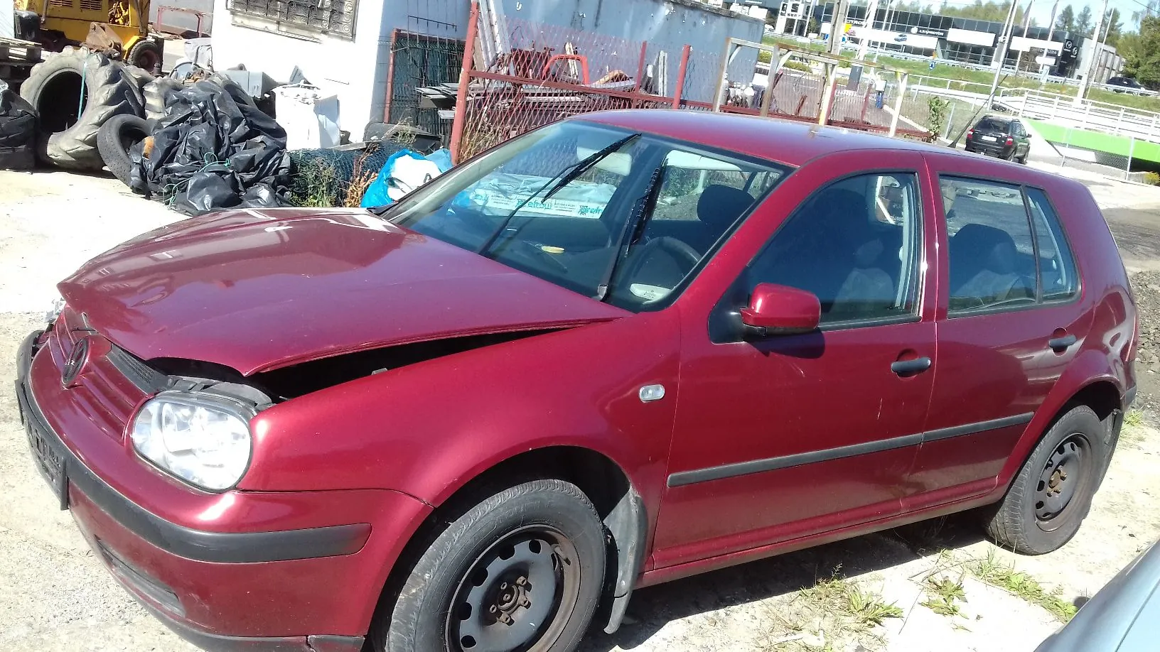 Uszkodzony, bordowy samochód osobowy marki Volkswagen Golf zaparkowany na placu. Pojazd ma wyraźne wgniecenie maski silnika w przedniej części. Auto stoi bokiem do obiektywu, w tle widoczne są elementy składowiska części samochodowych, opony oraz fragmenty budynków.