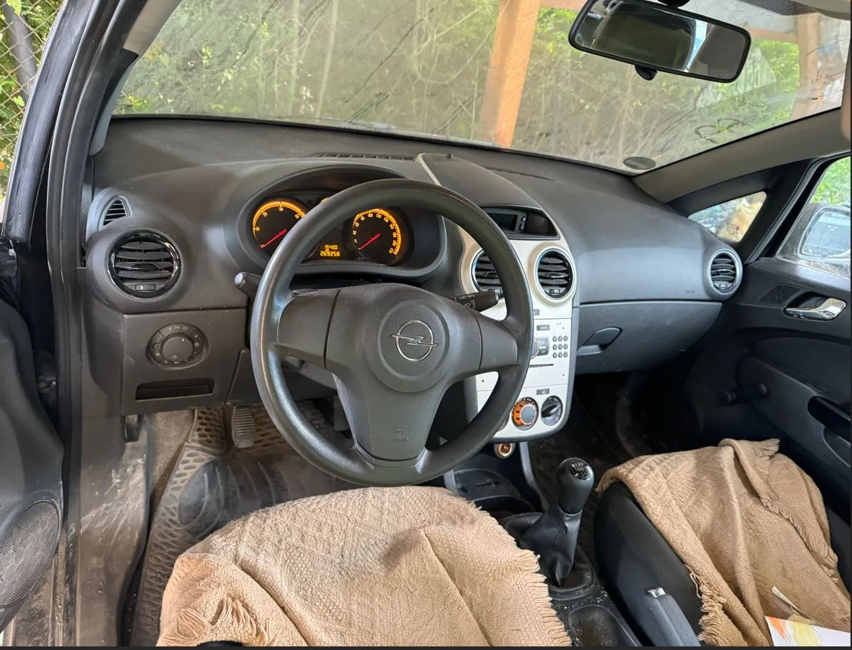 Wnętrze samochodu marki Opel widziane z perspektywy kierowcy. Widoczna jest czarna kierownica z logo producenta, deska rozdzielcza z analogowymi wskaźnikami prędkości i obrotów oraz centralna konsola z przyciskami i pokrętłami klimatyzacji. Fotele są przykryte beżowymi, teksturowanymi pokrowcami lub tkaninami. Przez przednią szybę widoczna jest zieleń drzew.