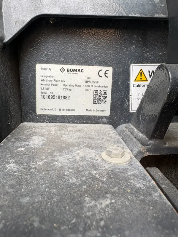 Zbliżenie na tabliczkę znamionową maszyny marki BOMAG. Na białej etykiecie widnieją następujące informacje: 'Made by BOMAG', 'Designation: Vibratory Plate rev.', 'Type: BPR 35/60', 'Nominal Power: 3,6 kW', 'Operating Mass: 205 kg', 'Year of Construction: 2021', 'Serial-No.: 101695181882'. Widoczny jest również znak CE, kod QR oraz napis 'Hellerwald, D-56154 Boppard Made in Germany'. Tabliczka jest przytwierdzona do czarnej, metalowej obudowy urządzenia.