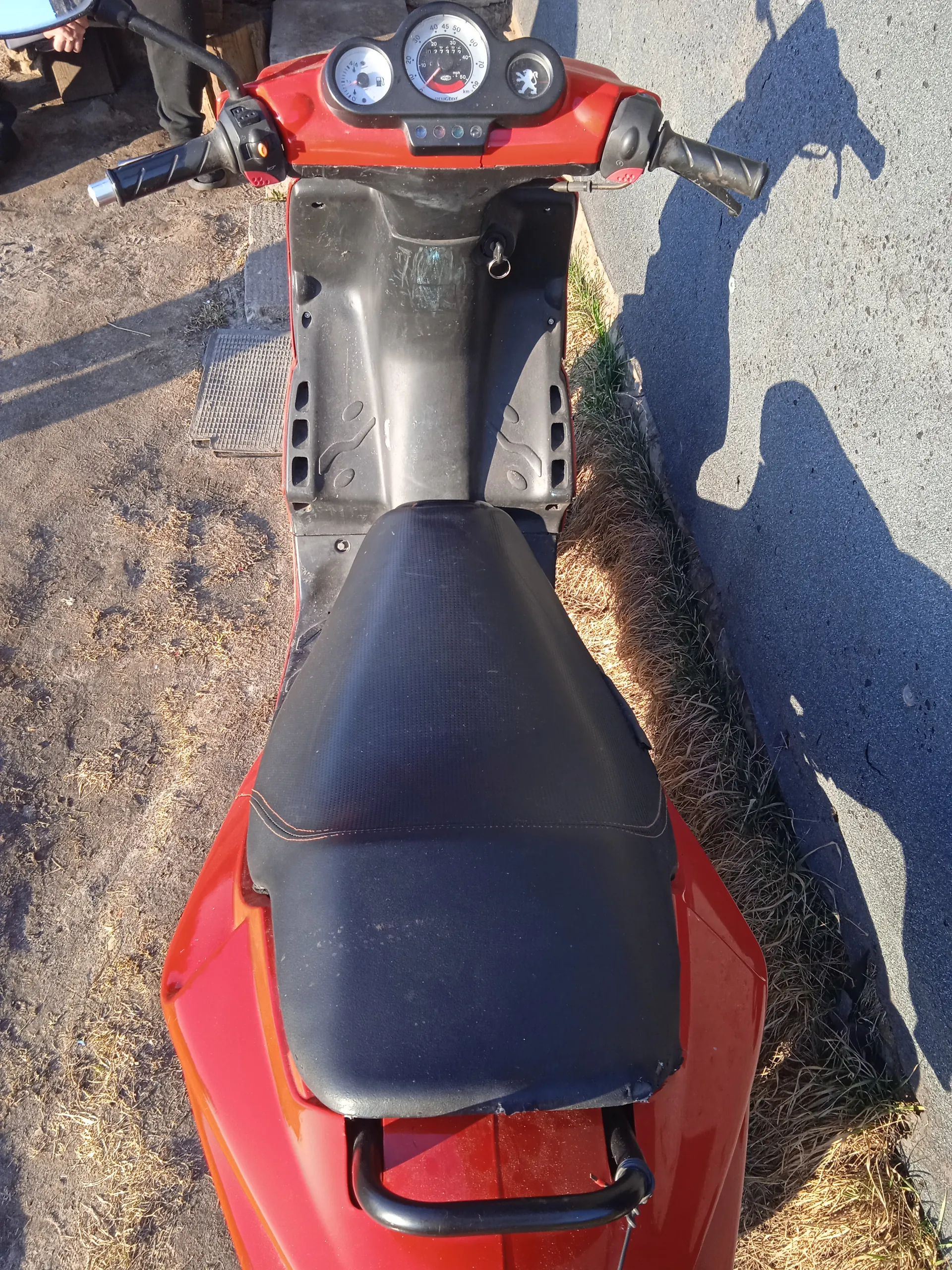 Czerwony skuter widziany z góry. Czarna siedziba, kierownica z trzema wskaźnikami (środkowy z licznikiem prędkości, prawy z logo Peugeot), czerwone nadwozie. Tło: asfalt, sucha trawa, ziemia oraz cień osoby na prawo od skutera. W tle widoczne nogi stojącej osoby i metalowa kratka.