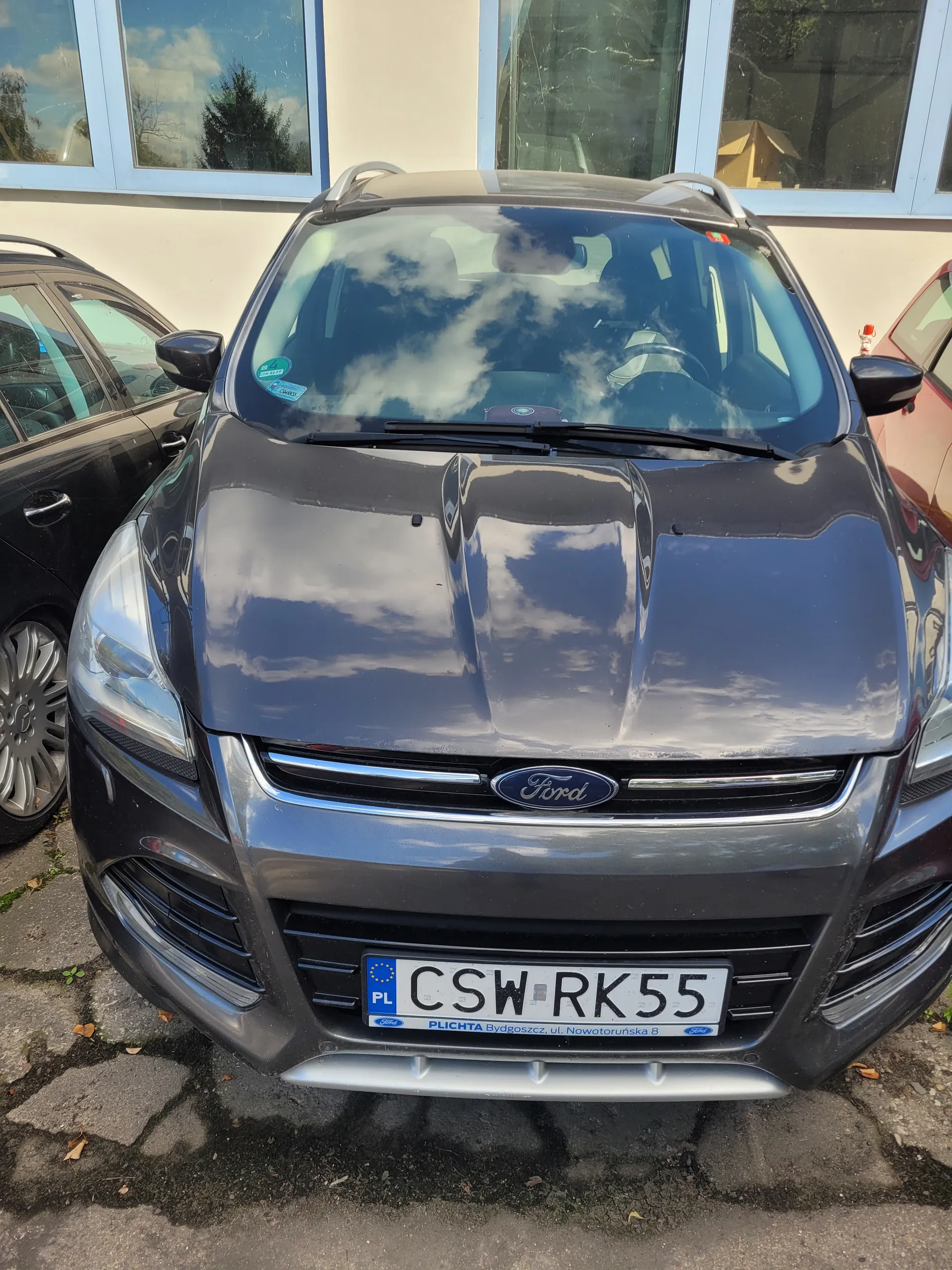 Ciemnoszary samochód marki Ford z przodu, z widocznym logo na atrapie chłodnicy. Tablica rejestracyjna CSW RK55 z napisem PŁOCHTA Bydgoszcz ul. Nowotoruńska 8 oraz oznaczeniem PL i symbolem Unii Europejskiej. Na tle budynek z niebieskimi ramami okiennymi odbijającymi niebo i drzewa, oraz fragment innego pojazdu. Asfaltowa powierzchnia z widocznymi pęknięciami.