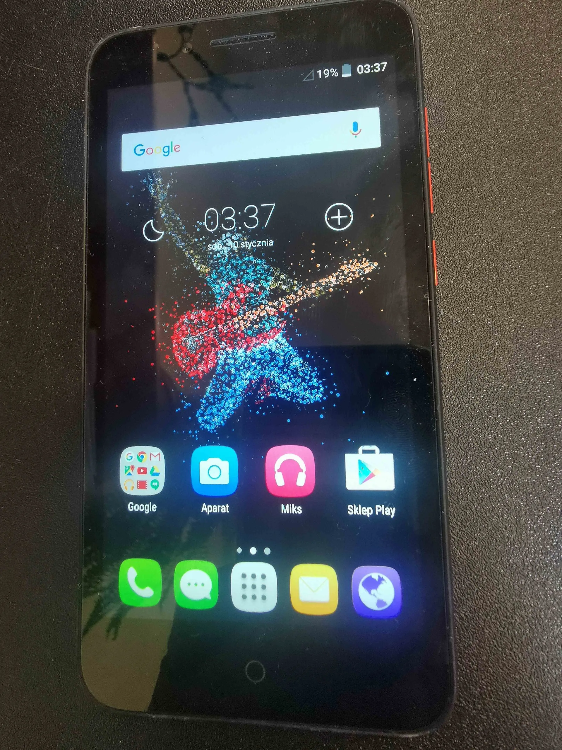 Czarny smartfon z włączonym ekranem głównym. Na górze znajduje się pasek stanu z ikoną baterii 19% i godziną 03:37. Poniżej widoczny jest pasek wyszukiwania Google. Na środku ekranu znajduje się duży zegar cyfrowy wskazujący 03:37 oraz data '30 stycznia'. Tło ekranu przedstawia artystyczną grafikę postaci grającej na gitarze, złożoną z kolorowych punktów. W dolnej części ekranu widoczne są ikony aplikacji: Google, Aparat, Miks, Sklep Play oraz w najniższym rzędzie ikony telefonu, wiadomości, menu, poczty i przeglądarki.