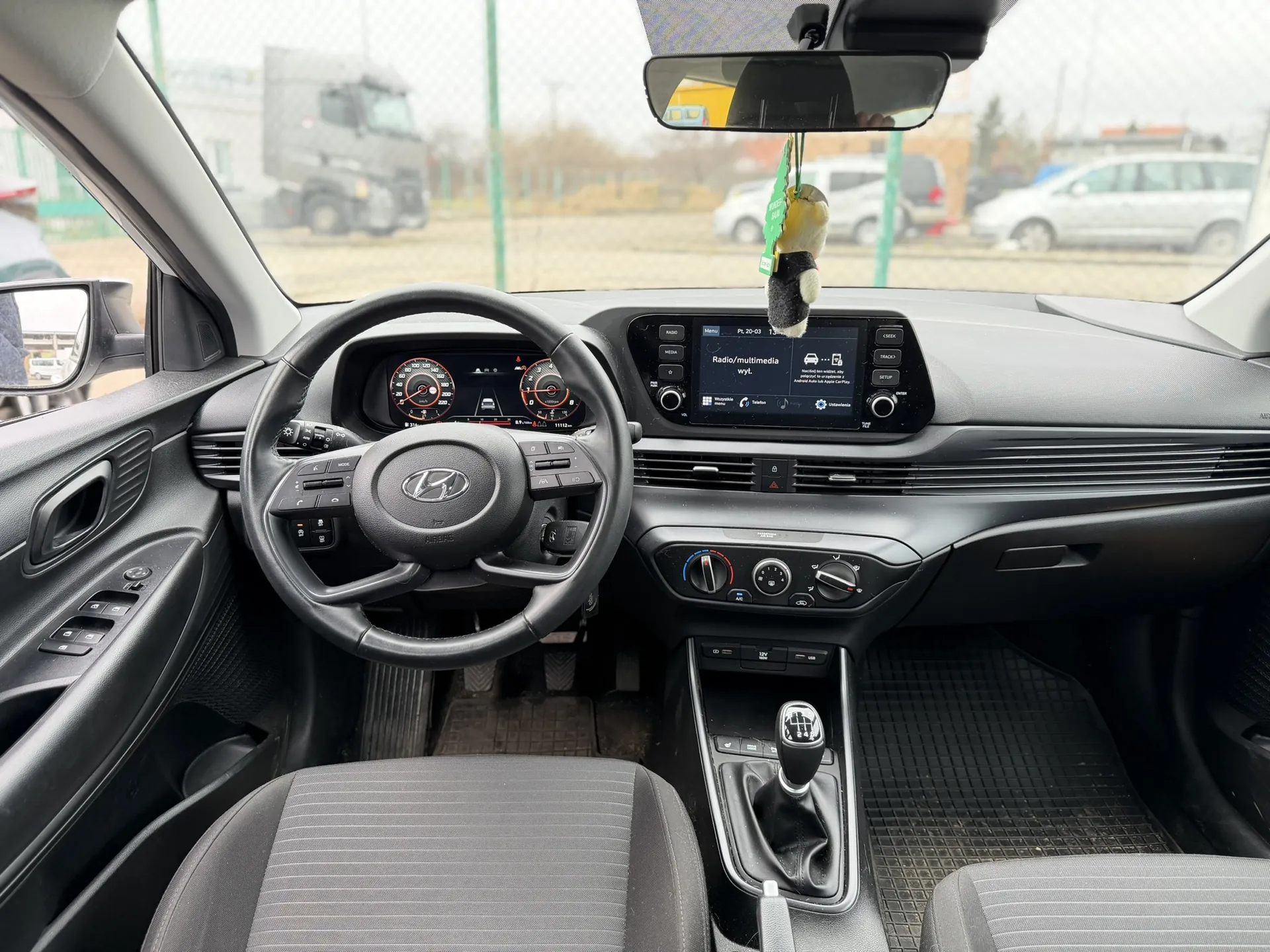 Wnętrze samochodu marki Hyundai z widocznym kierownicą z logo producenta, tablicą przyrządów z czerwonymi wskaźnikami, ekranem multimedialnym wyświetlającym napis "Radio/multimedia wyl.", manualną skrzynią biegów, szarymi siedzeniami w paski oraz lusterkiem wstecznym z wiszącą dekoracją w kształcie pingwina. Przez przednią szybę widoczne są inne pojazdy, ogrodzenie i budynki na zewnątrz.