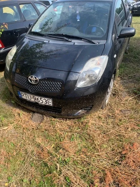 Czarny samochód marki Toyota zaparkowany na trawiastym i piaszczystym podłożu. Widoczny jest przód pojazdu z tablicą rejestracyjną o numerze PTU 9653E. Samochód jest zakurzony, a obok niego stoi inny czarny pojazd.