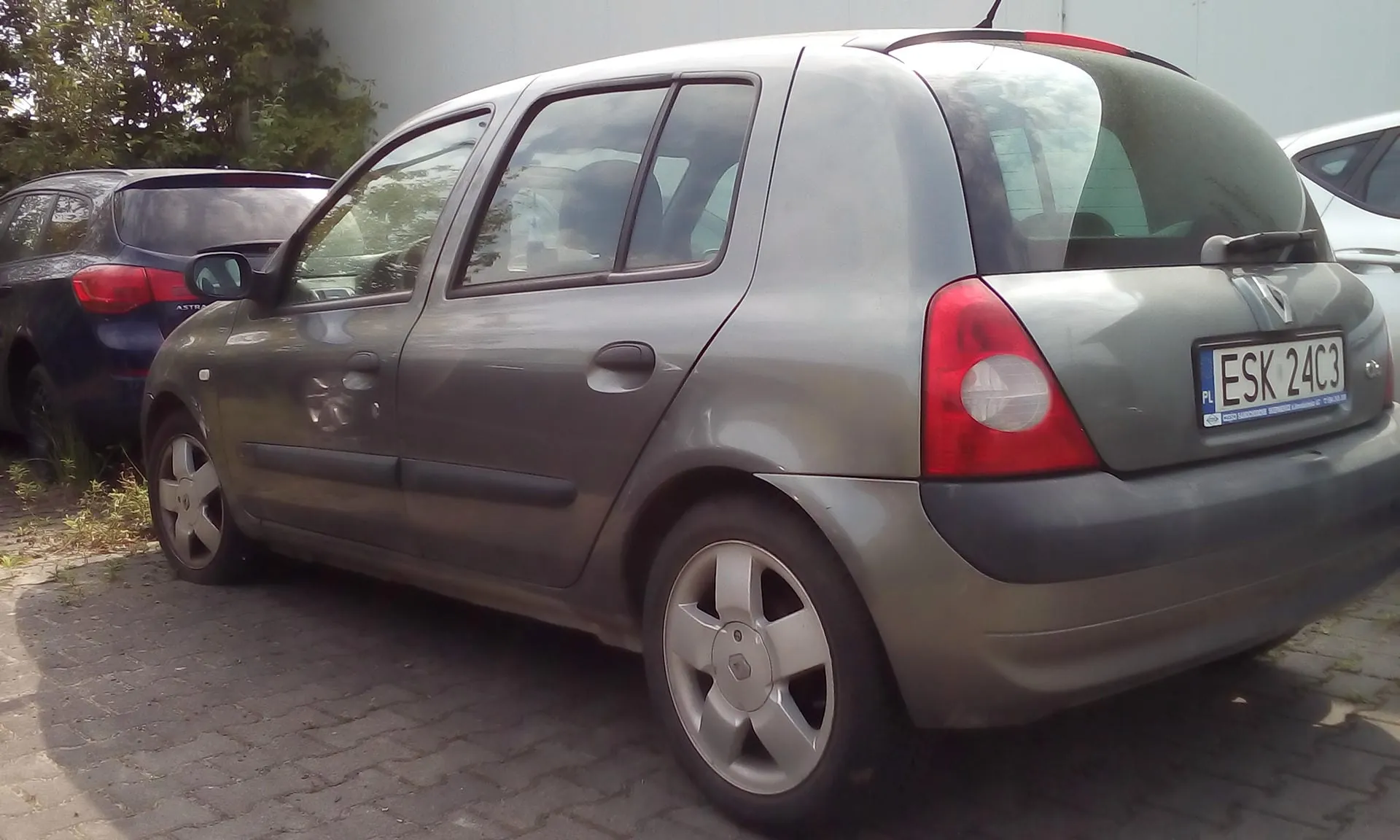 Srebrny samochód osobowy marki Renault Clio zaparkowany na kostce brukowej. Widok z tyłu i z boku pojazdu. Samochód posiada polską tablicę rejestracyjną o numerze ESK 24C3. W tle widoczny jest fragment innego, ciemnoniebieskiego samochodu oraz zielone drzewa.