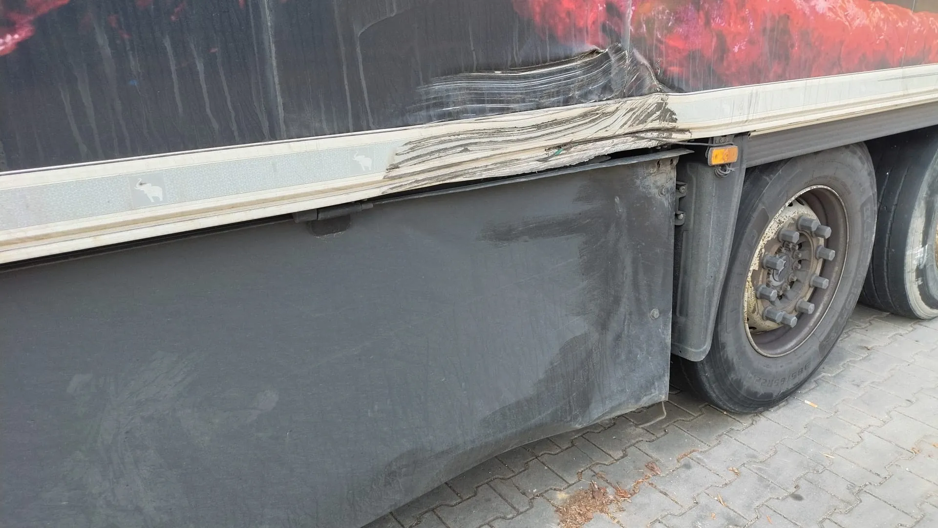 Zbliżenie na uszkodzoną część boczną ciężarówki. Widoczne są zgięte metalowe elementy nad kołem, czarna osłona koła oraz fragment nadwozia z czerwonym wzorem. Na srebrnej listwie biegnącej wzdłuż nadwozia znajdują się małe ikony słoni. Koło z rdzewiejącym felgiem i śrubami, a także oponą z widocznymi znakami zużycia. Podłoga wyłożona jest kostką brukową. Brak czytelnego tekstu.