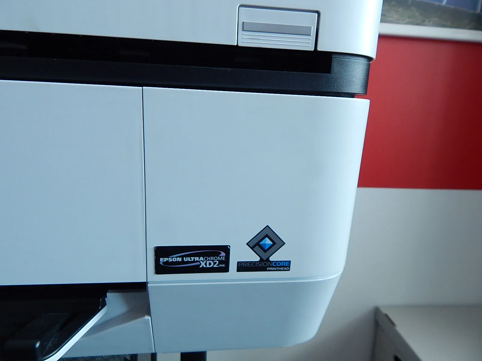 Zbliżenie na białą obudowę drukarki Epson. Na przednim panelu widoczne są dwie naklejki: jedna z napisem „EPSON ULTRACHROME XD2 INK” oraz druga z logo w kształcie rombu i napisem „PRECISIONCORE PRINTHEAD”. W górnej części obrazu widoczny jest fragment tacy papieru oraz czarna szczelina wyjściowa.