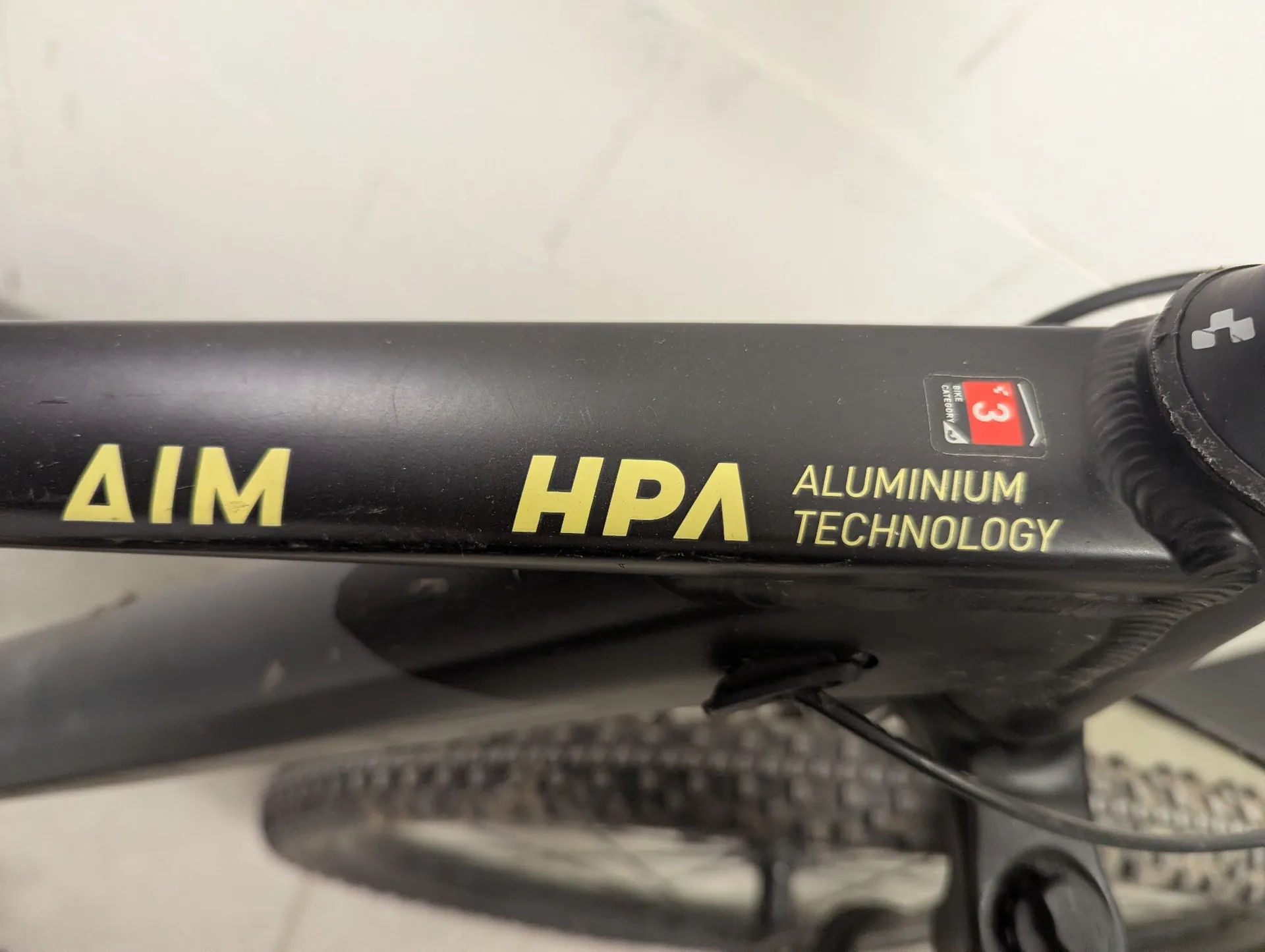 Zbliżenie na czarną ramę roweru z żółtymi napisami „AIM HPA” oraz białym napisem „ALUMINIUM TECHNOLOGY”. Obok znajduje się mała czerwono-biała naklejka z cyfrą 3 i napisem „Made in Germany”. W tle widoczny jest fragment opony rowerowej oraz biała ściana.